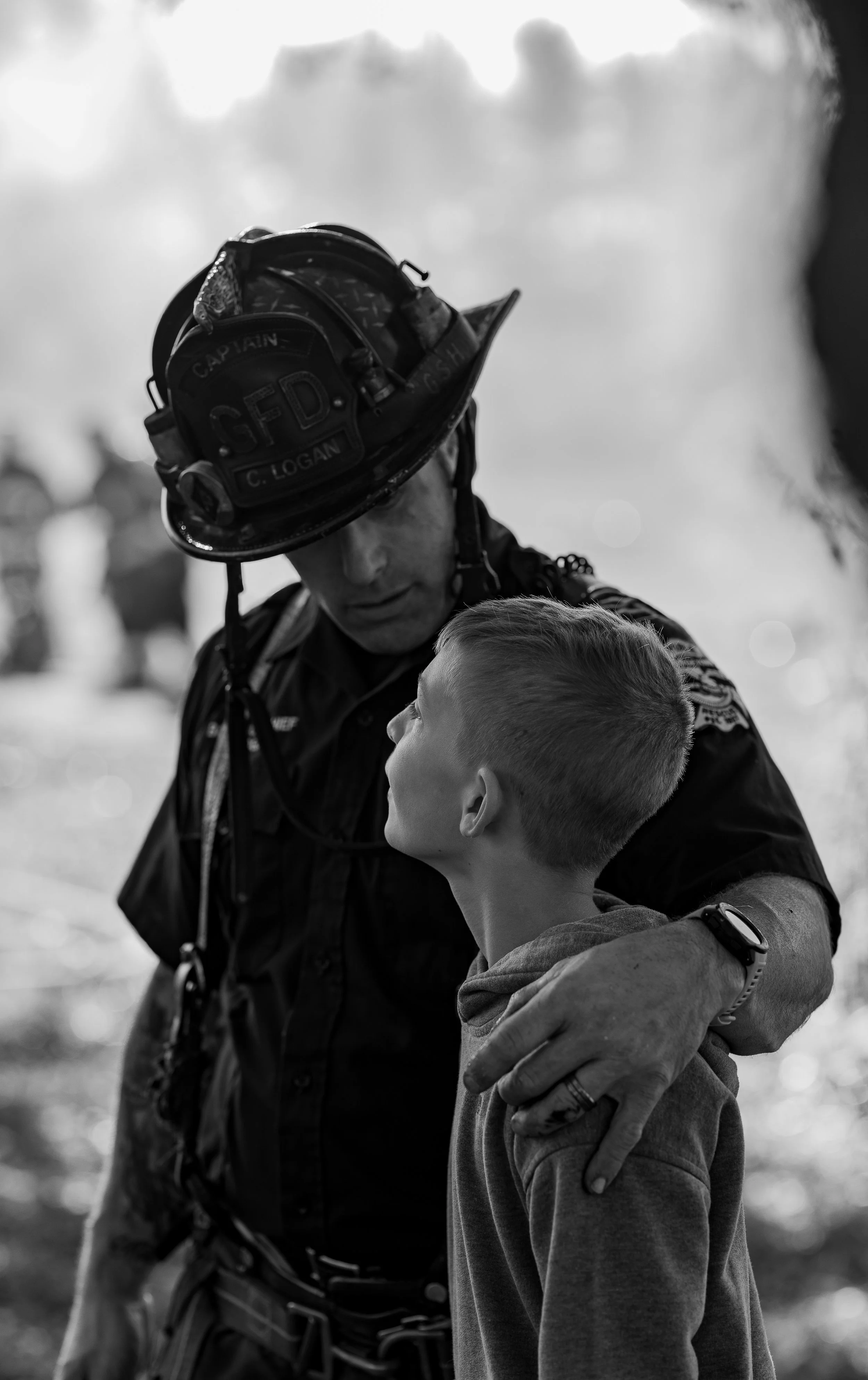 A firefighter in uniform and helmet comforting a young boy outdoors in black and white.