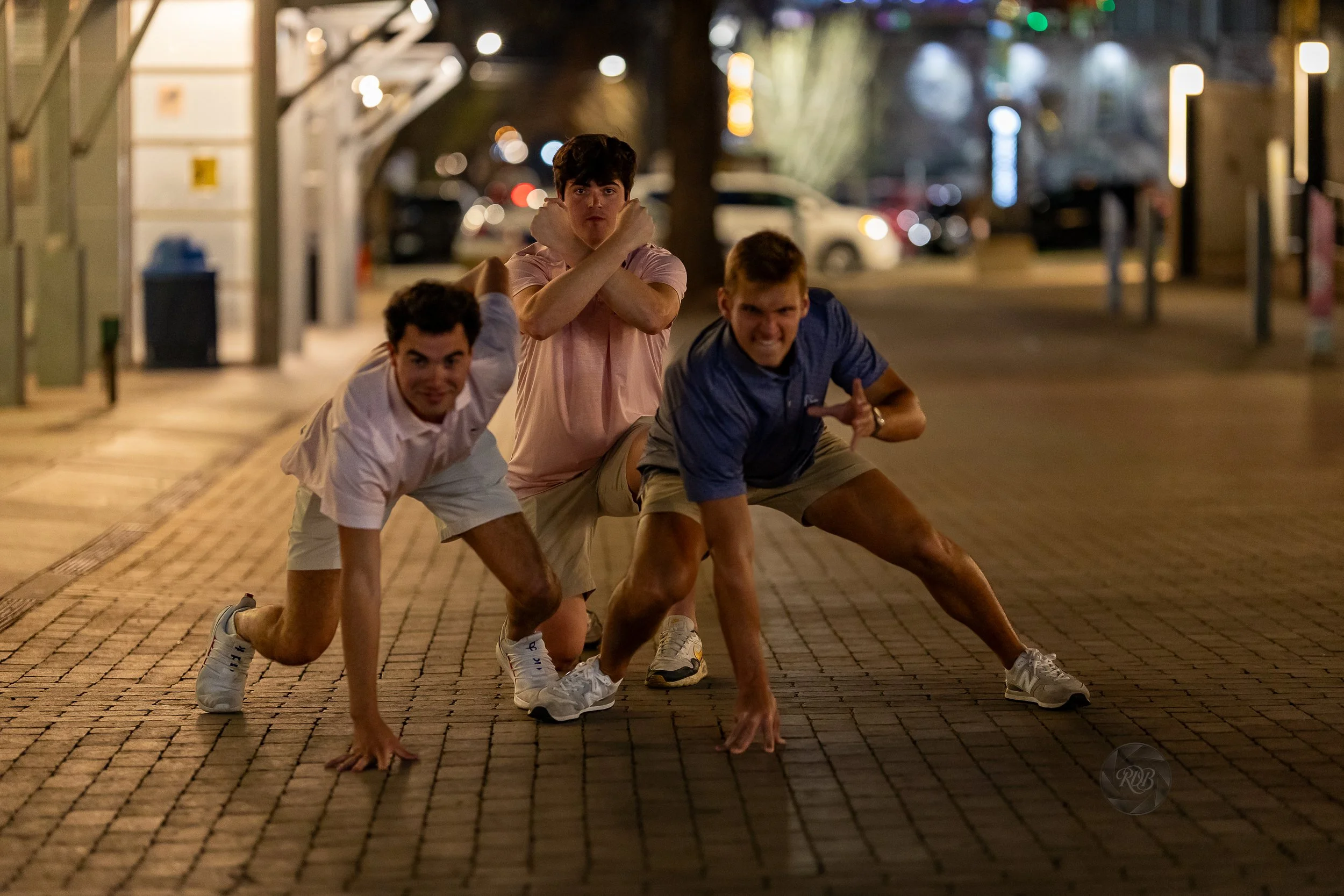 Three young men posing playfully on a brick-paved street at night, crouching with one arm extended and one arm crossed in front of their faces.