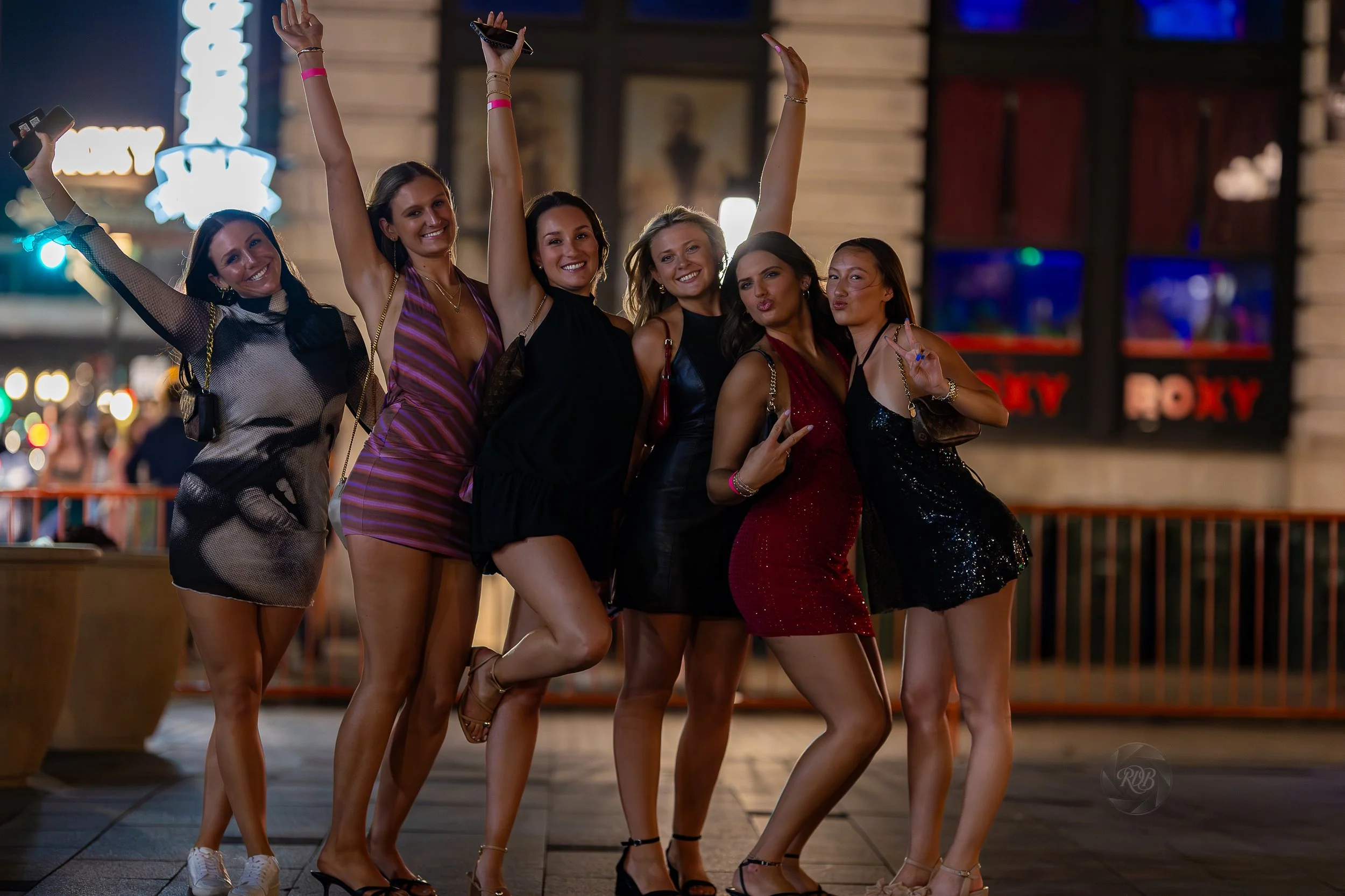 Group of six women celebrating at night, posing and smiling with some making peace signs, in an urban area with neon signs and lights in the background.