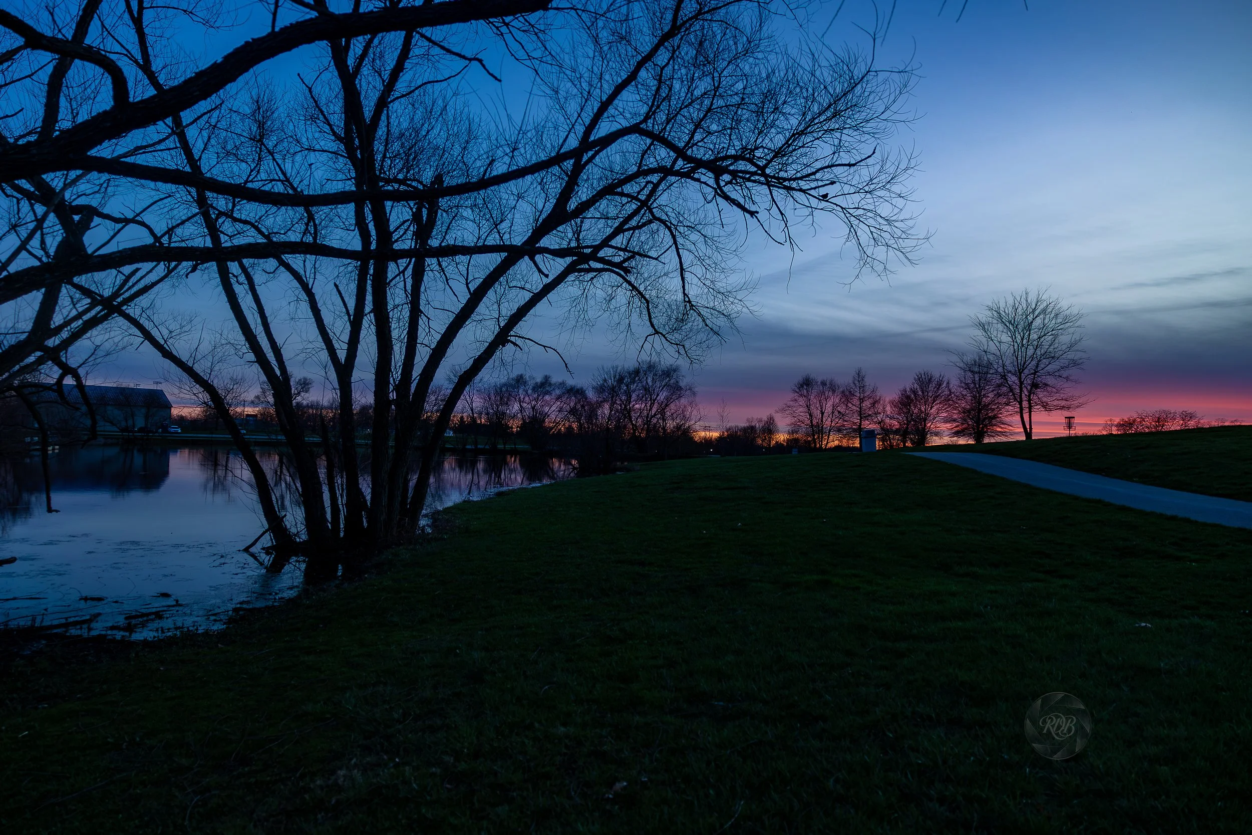A lakeside park scene at dusk with a leafless tree in the foreground, a calm water body reflecting the sky, and a paved pathway curving through a grassy area. The sky displays hues of pink, purple, and blue with sparse clouds.