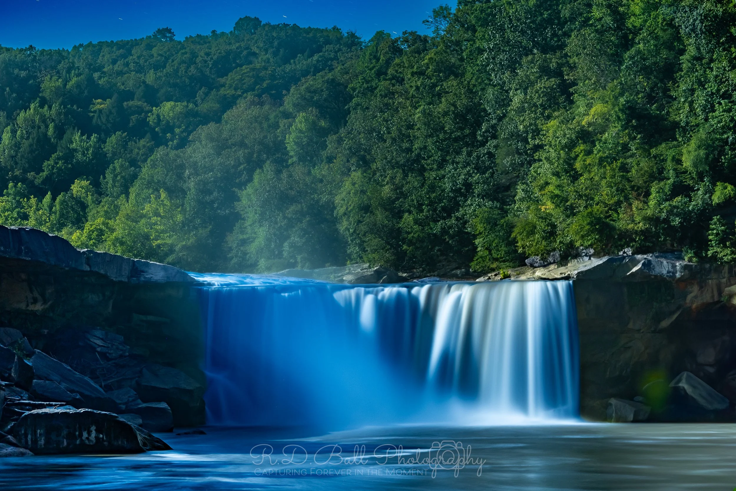 A cascading waterfall flowing into a river surrounded by lush green trees and forested hills under a blue sky.