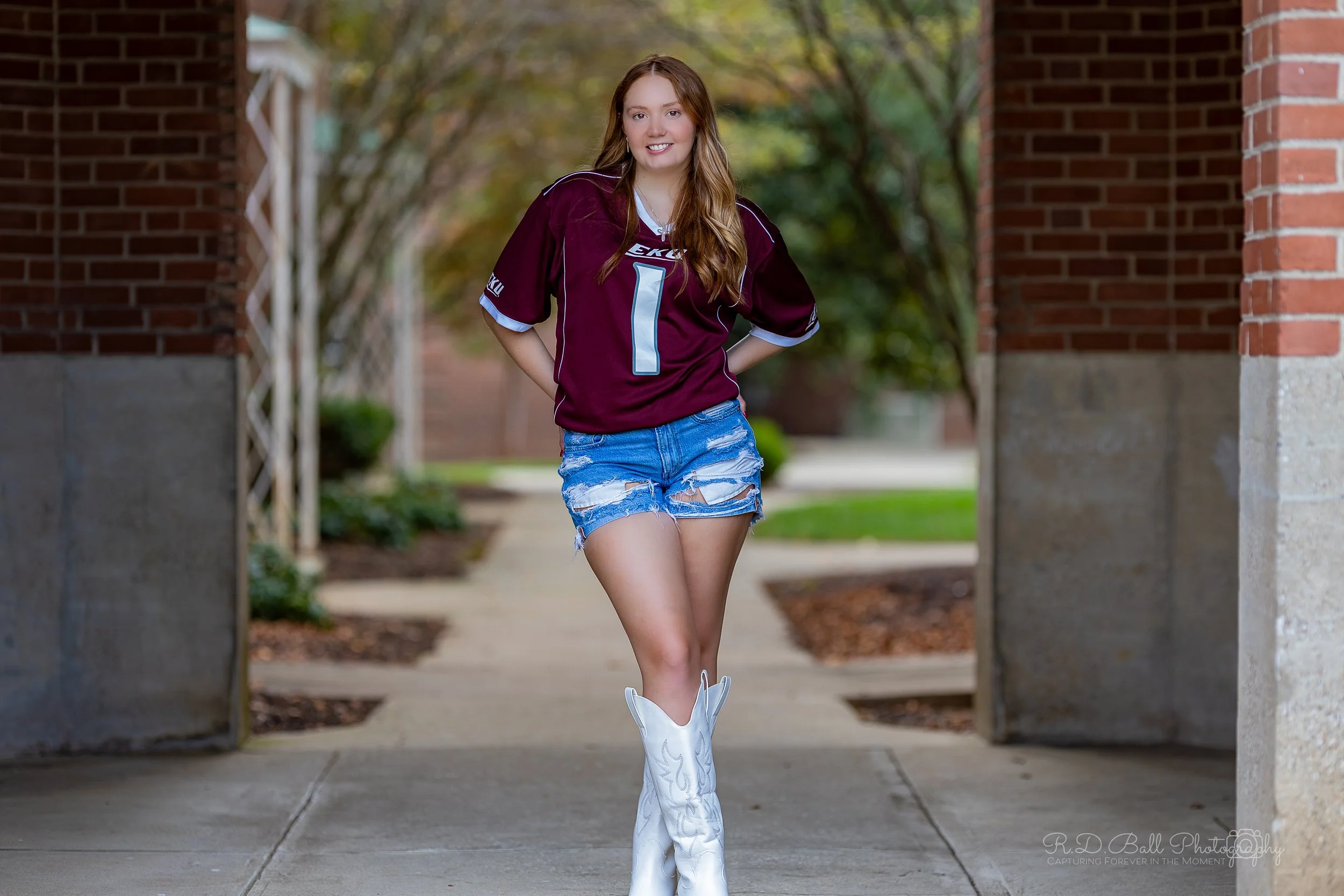 Young woman smiling outdoors at a college campus, wearing a maroon football jersey, ripped denim shorts, and white cowboy boots.