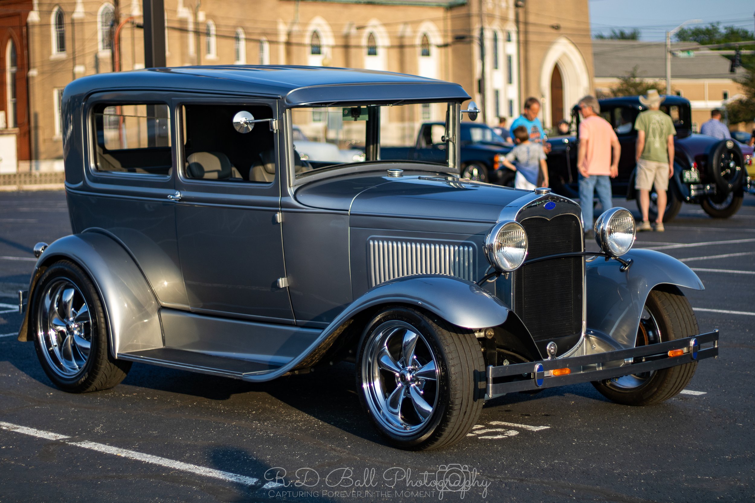 A vintage gray car parked in a lot during a classic car show with people in the background.
