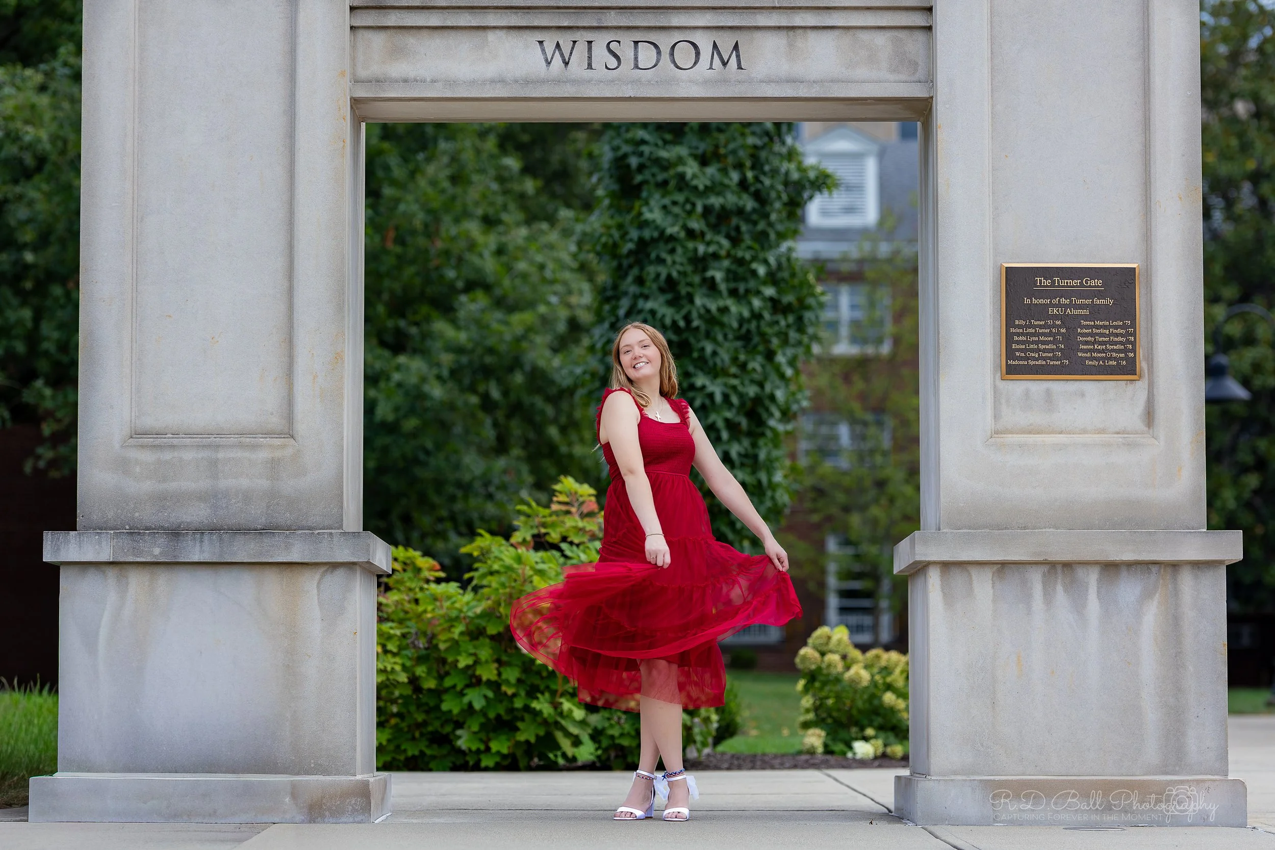 A young woman in a red dress standing in front of a stone archway with the word 'WISDOM' inscribed at the top. She is smiling, holding her dress, and wearing white heels, with greenery and a building in the background.