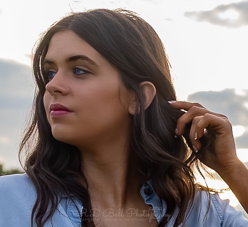 A young woman with dark wavy hair and blue eyes, wearing a light blue shirt, is touching her hair with her right hand. She is outdoors with a cloudy sky in the background.