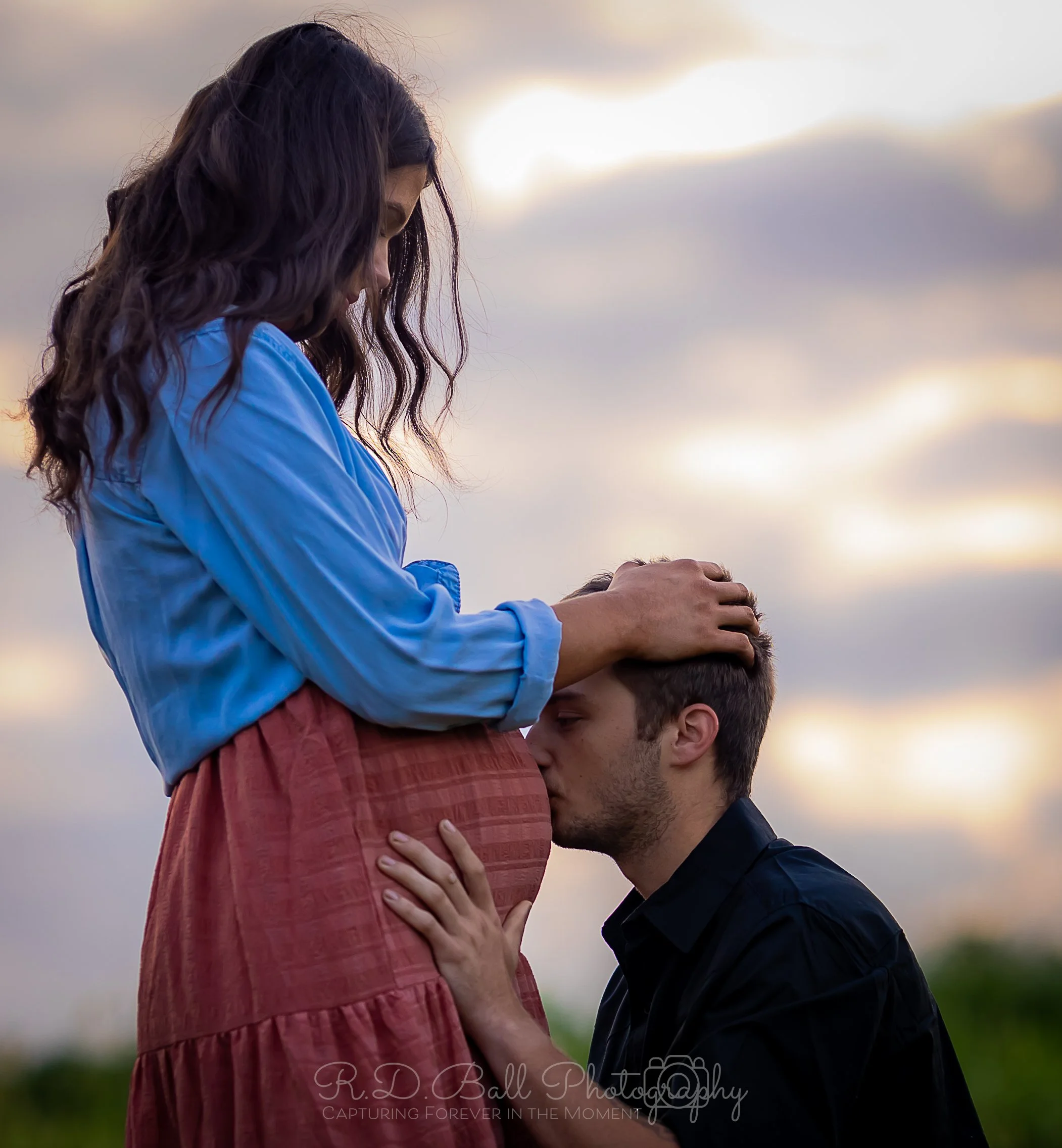 A man is kneeling and kissing a pregnant woman's belly while she gently touches his head. The woman is standing outdoors at sunset, wearing a blue blouse and a pink skirt, with an overcast sky in the background.