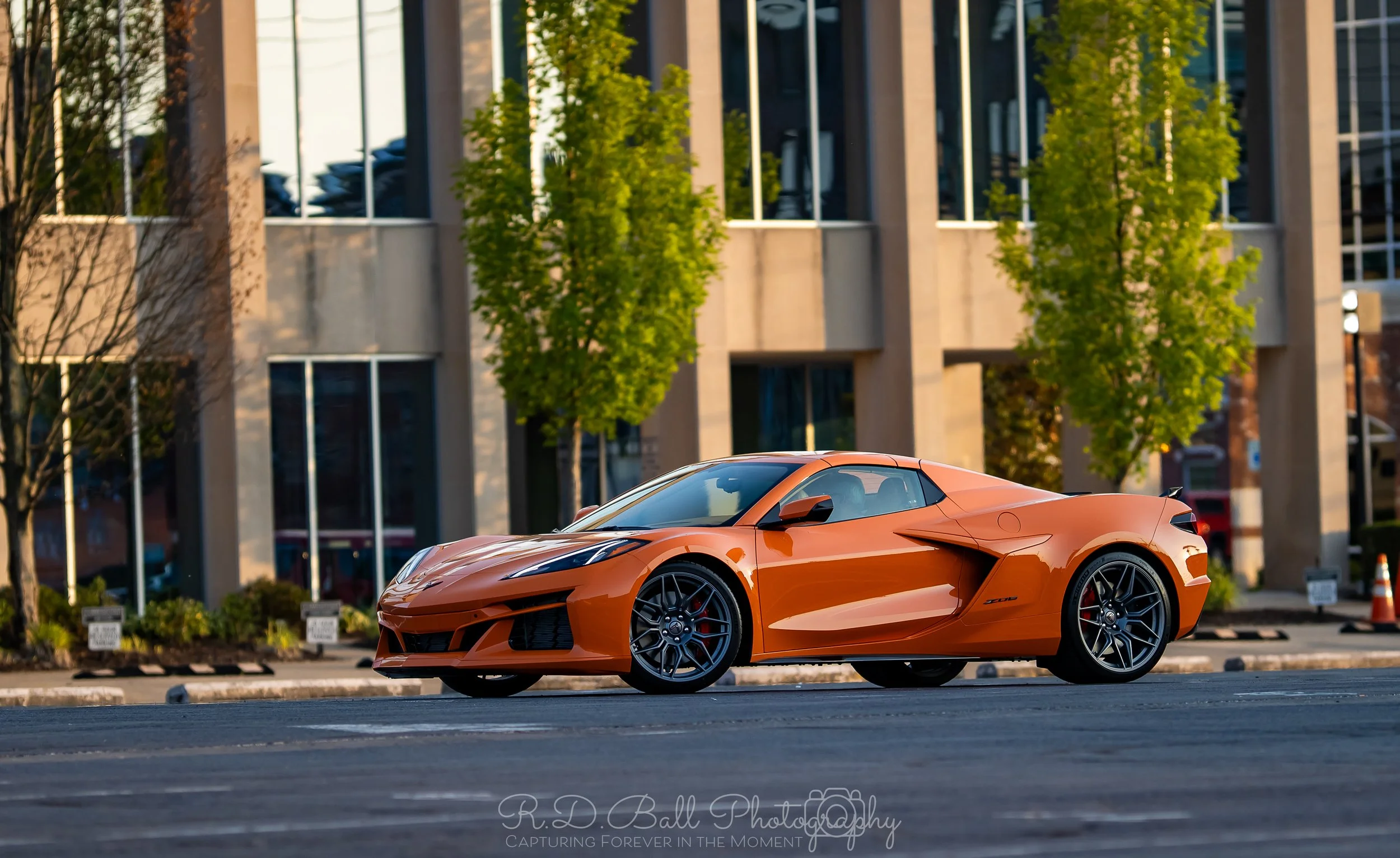 Orange sports car parked on city street with modern office building and green trees in background.