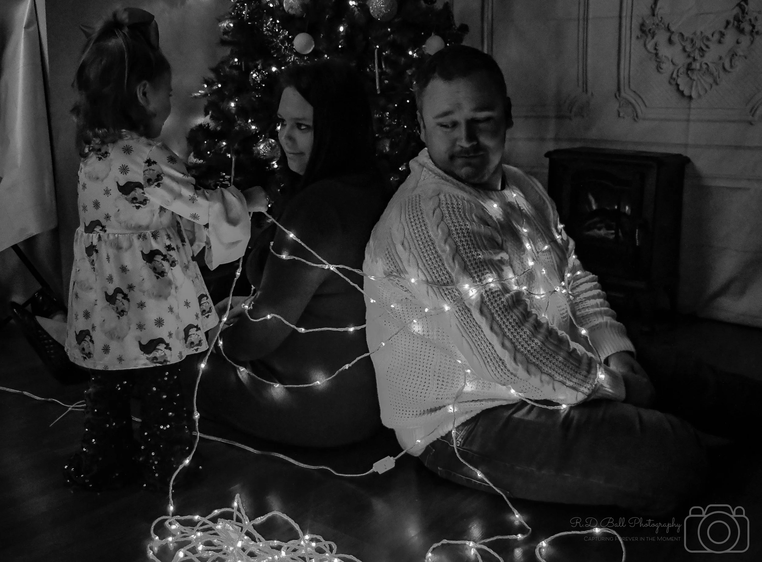 A family gathered around a decorated Christmas tree, with some members sitting and one standing, tangled in Christmas lights, in a cozy indoor setting.