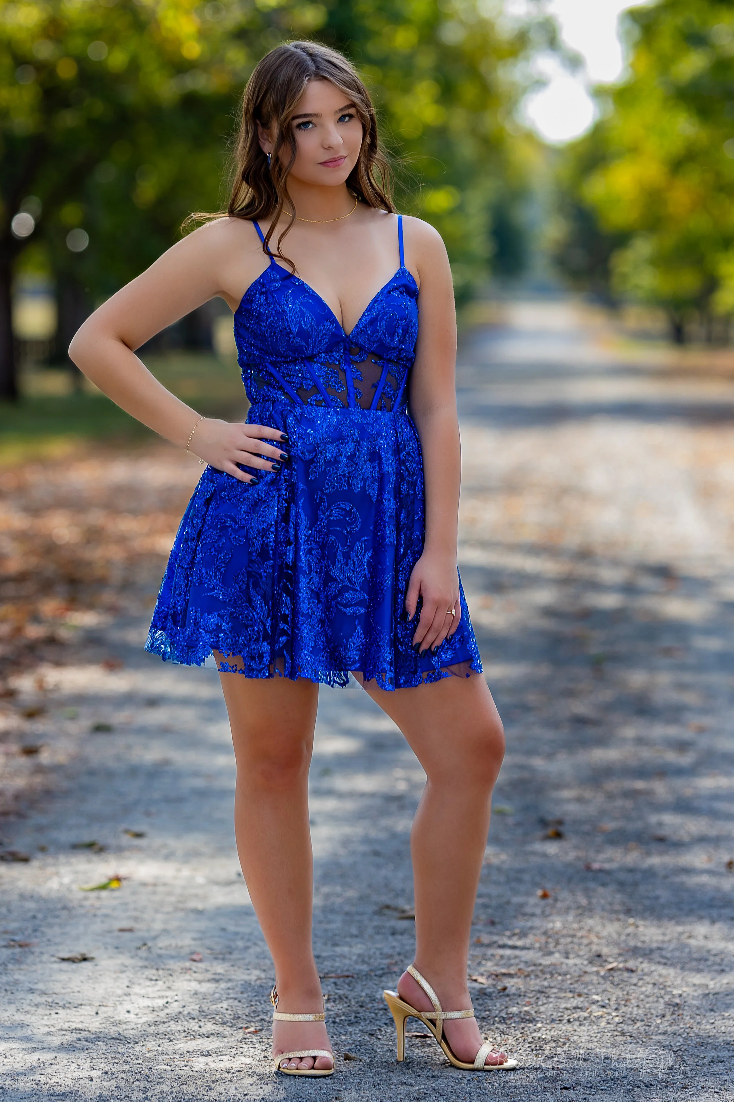 Young woman in a blue lace dress standing on a tree-lined dirt road in an outdoor setting.