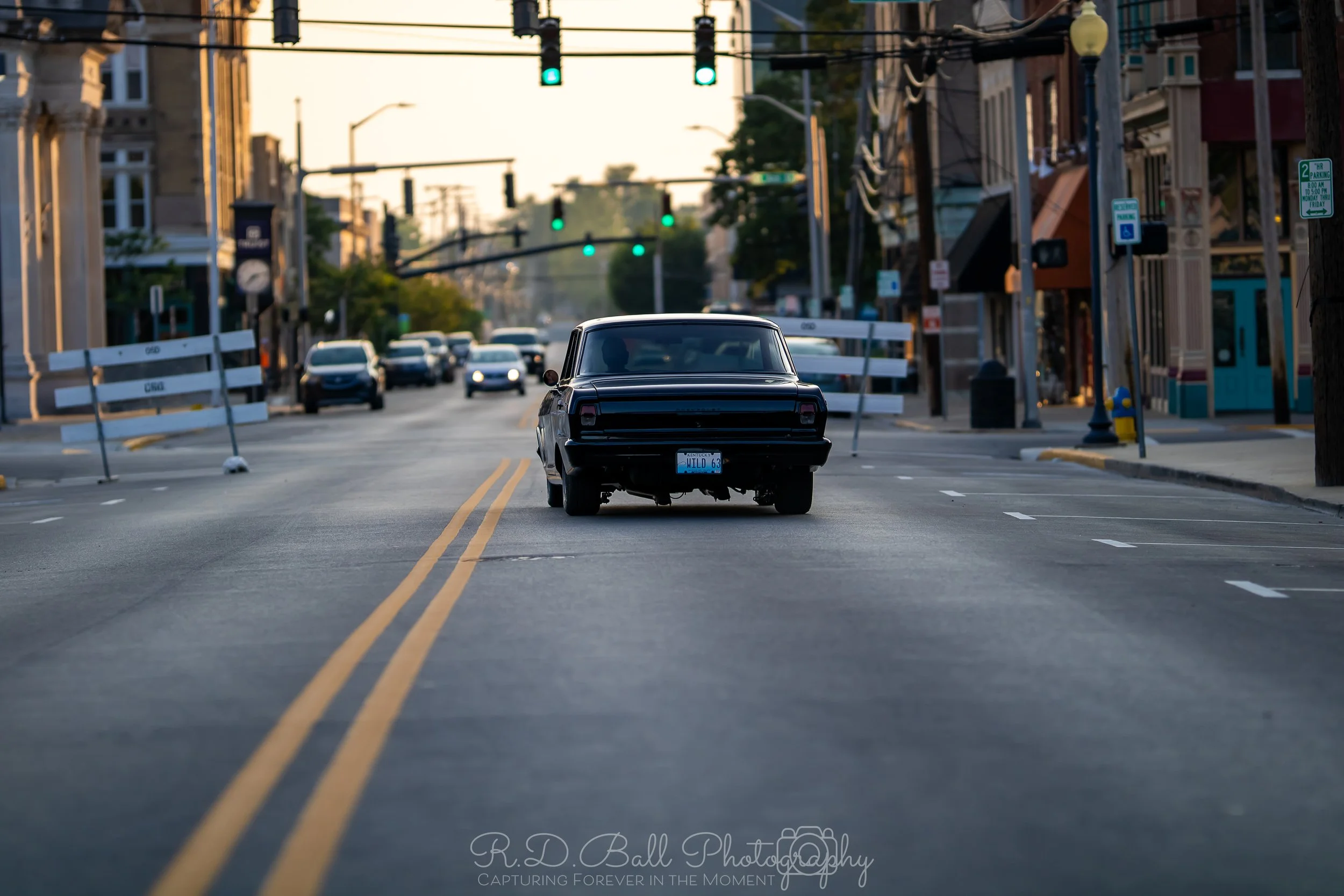 A black vintage car stopped in the middle of a city street with green traffic lights, surrounded by other cars and buildings on both sides.