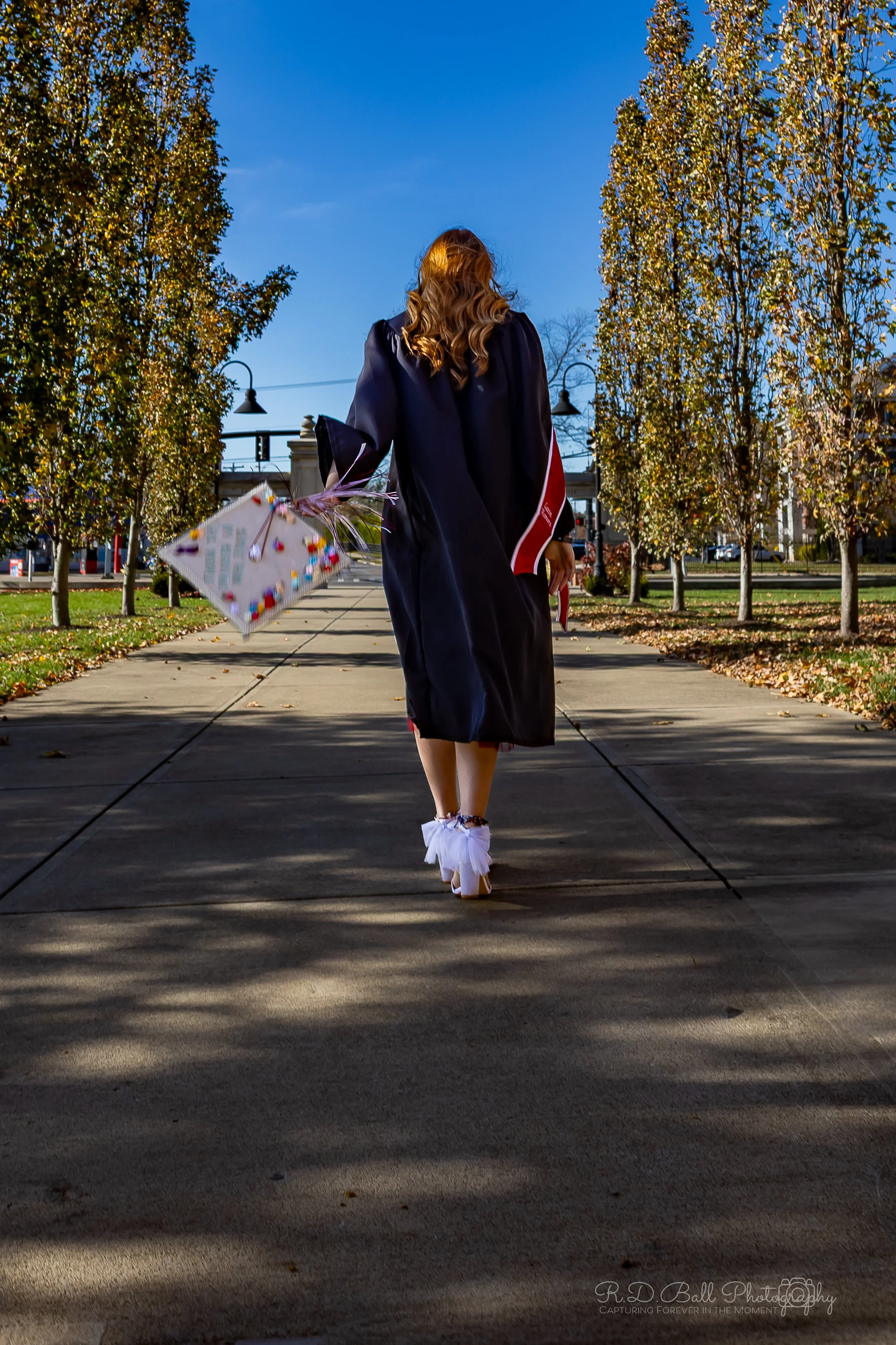 A woman dressed in a graduation cap and gown walking on a sidewalk carrying a decorated gift box with a bow, surrounded by autumn trees under a blue sky.