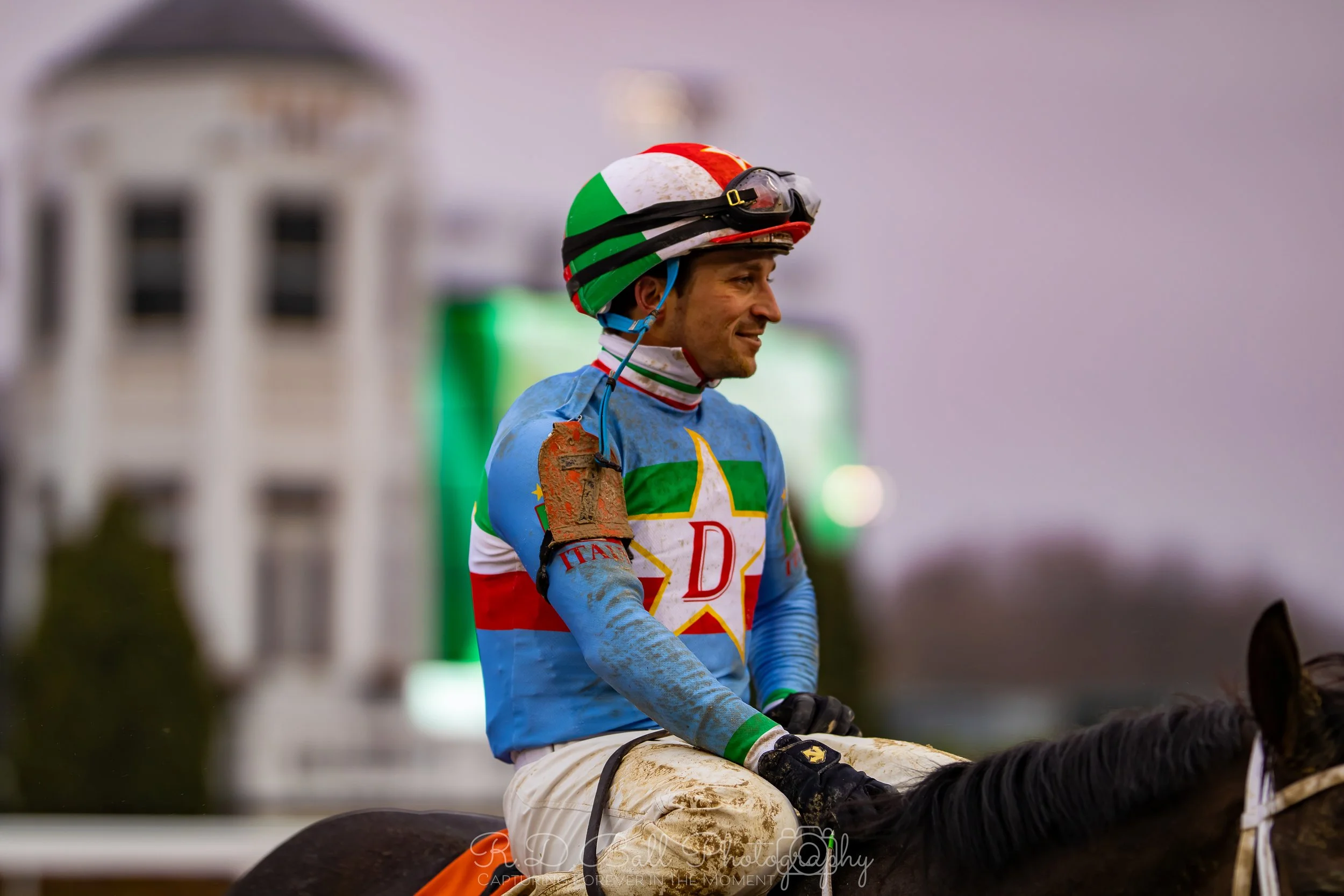 A jockey wearing colorful racing silks and a helmet, sitting on a muddy horse, with a blurred building in the background.