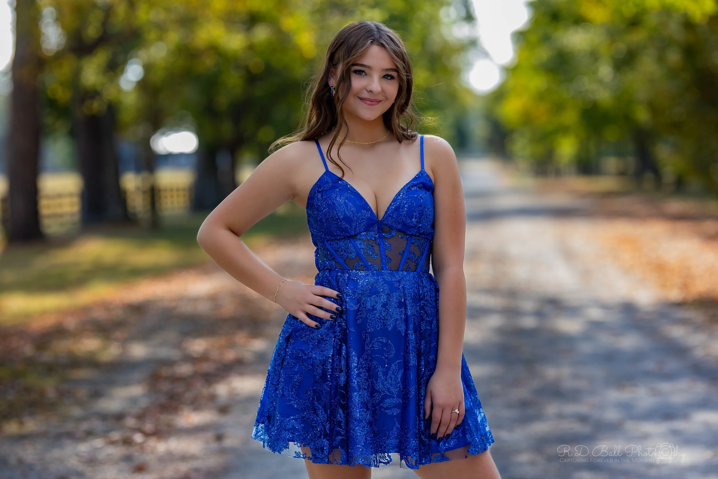A young woman in a blue lace dress standing outdoors on a dirt path with trees in the background.