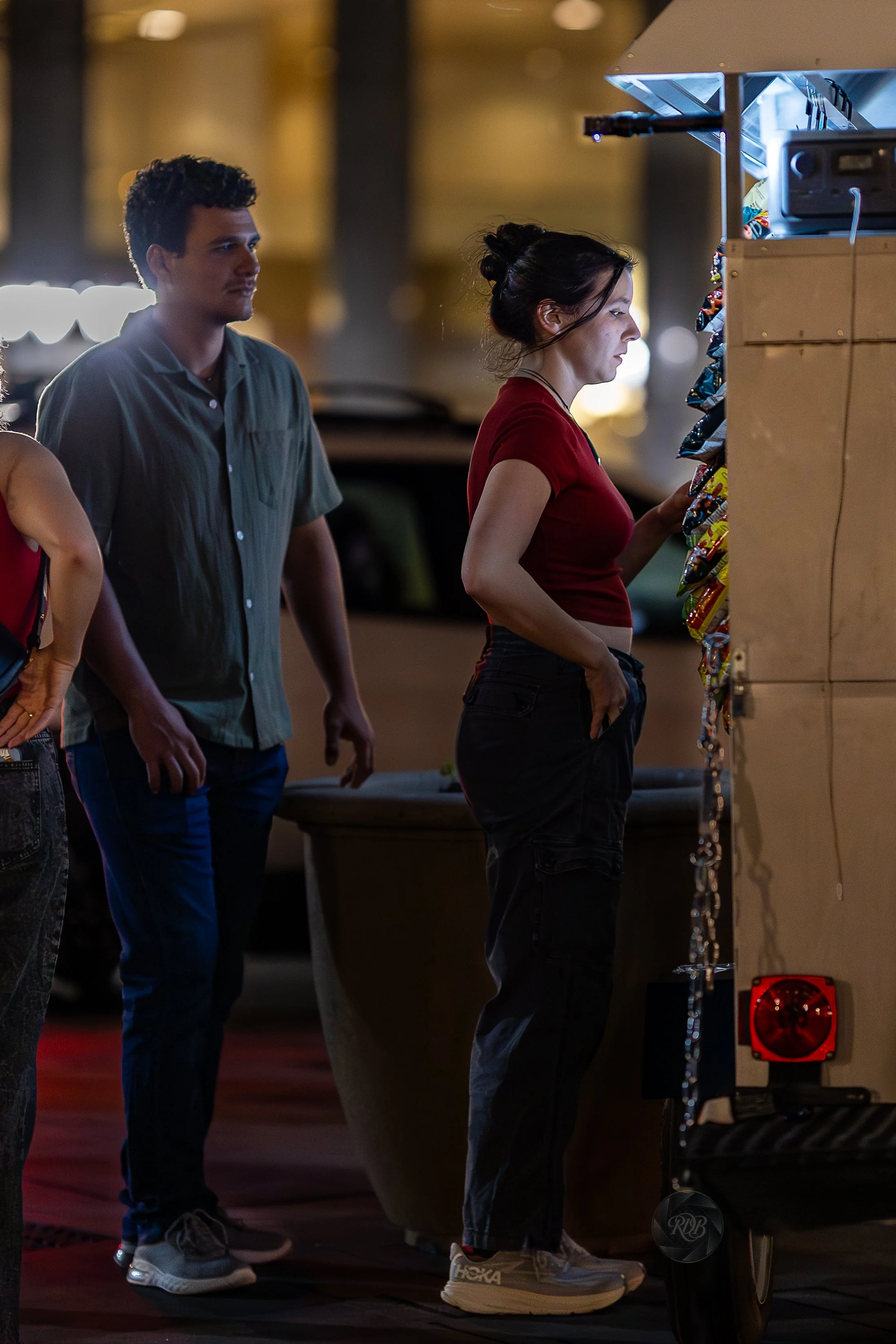 A woman in a red shirt and black cargo pants is looking at a vending machine at night, with a young man standing behind her.