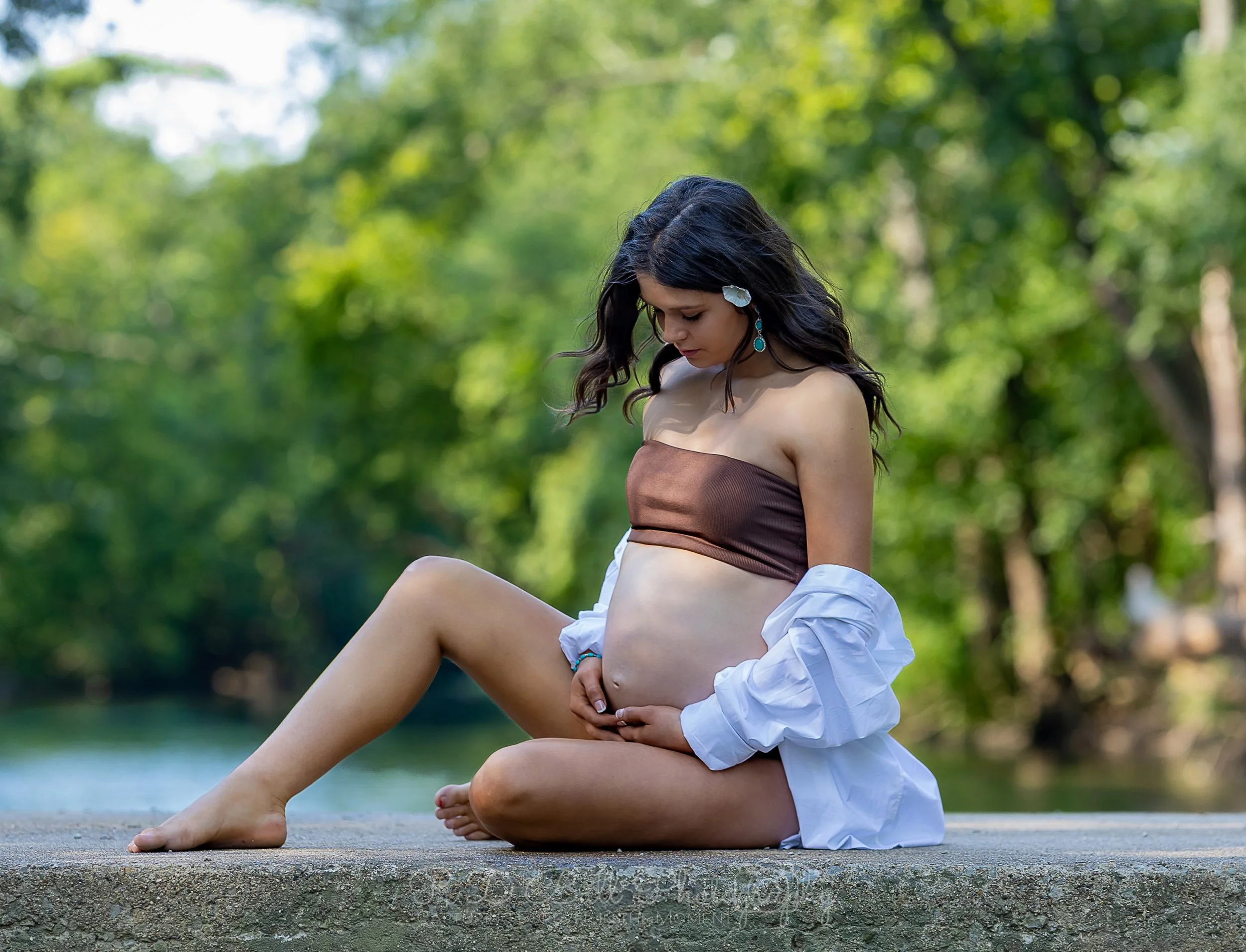 A pregnant woman sitting on a concrete surface outdoors, with green trees and water in the background. She is wearing a strapless brown top and a white shirt draped off her shoulders, holding her belly and looking down.