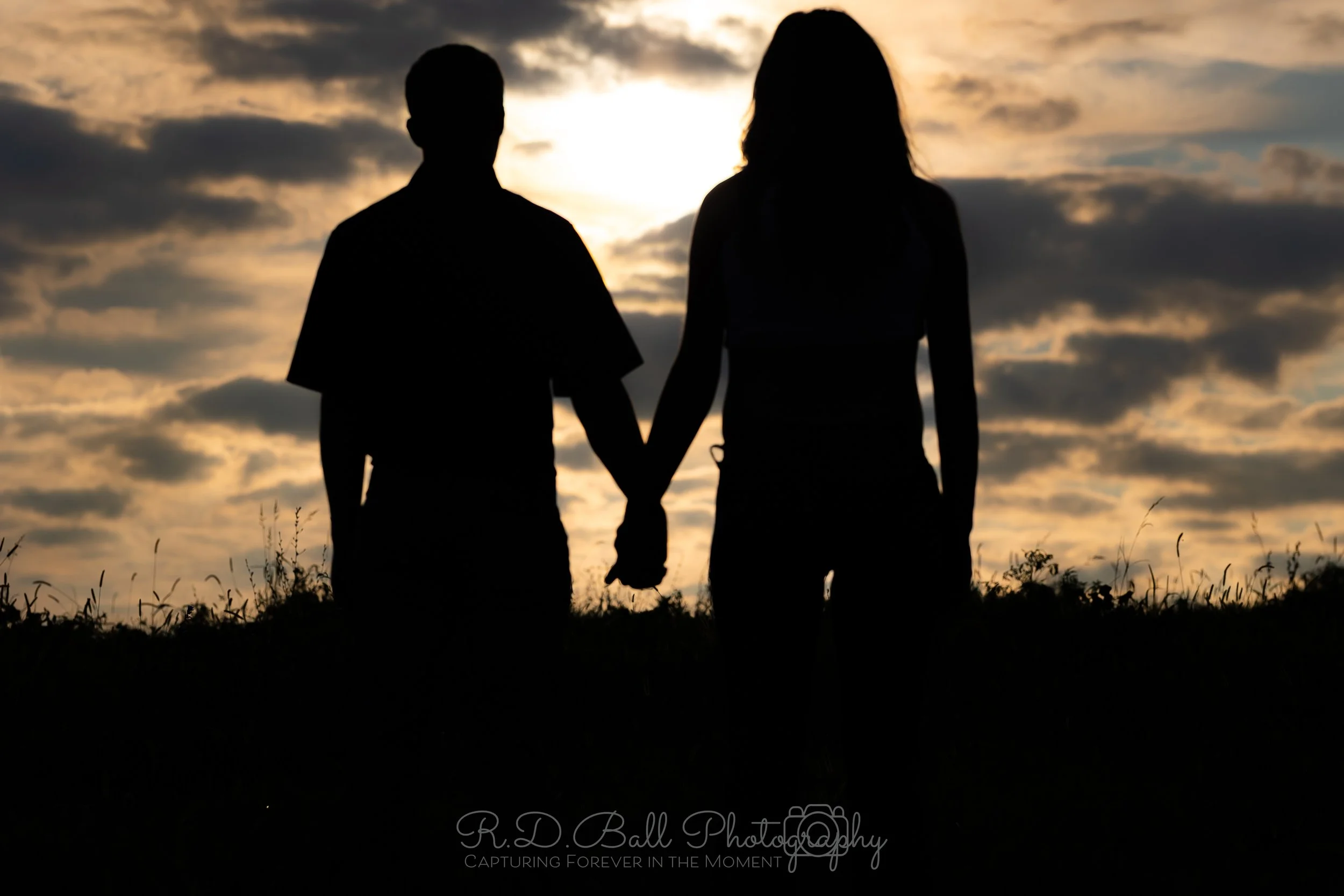 Silhouette of a couple holding hands on a hill during sunset with a cloudy sky in the background.