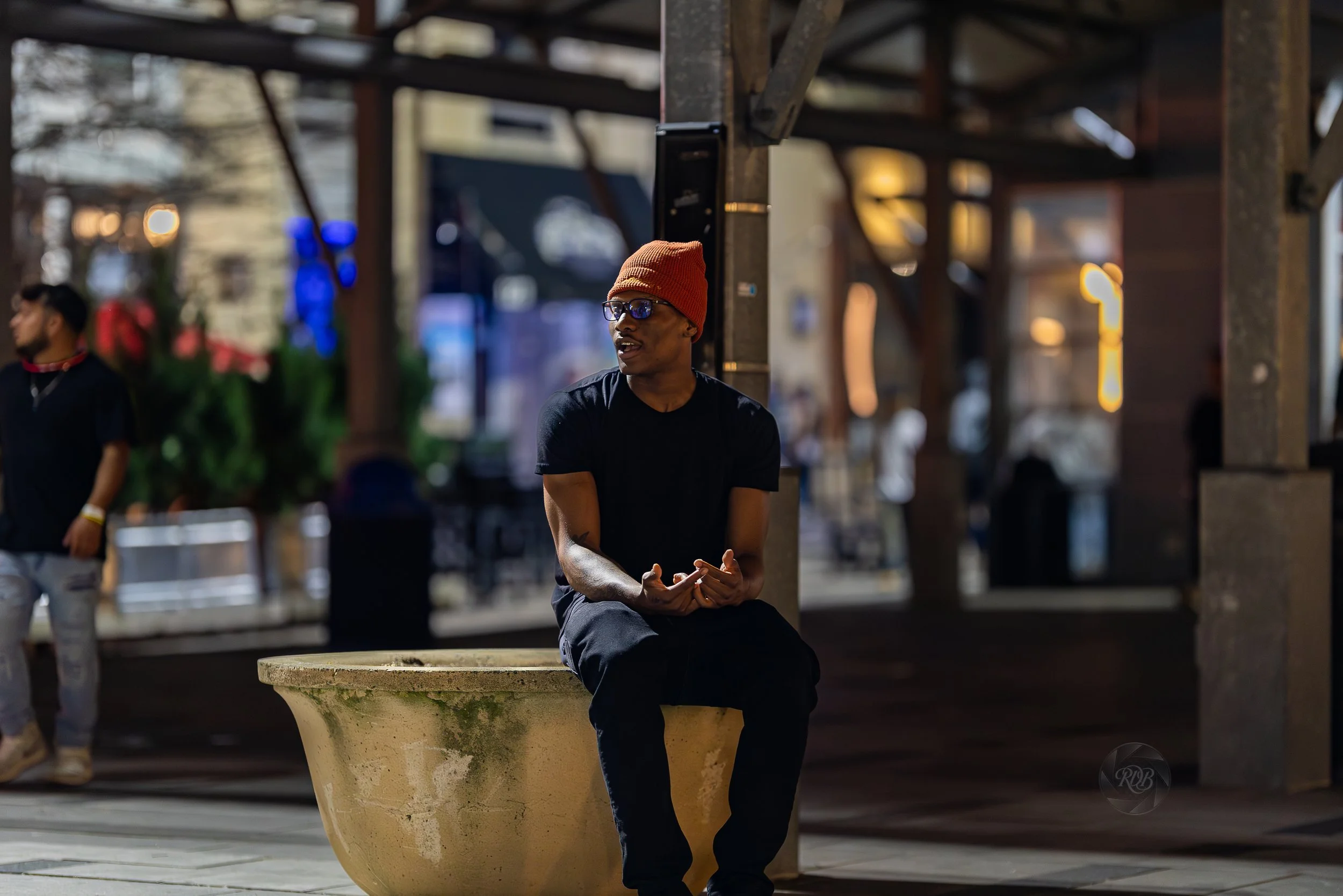 A man sitting on a large planter under a city pavilion at night, wearing glasses, a red beanie, a black t-shirt, and black pants, holding a phone in his hands. In the background, another person stands near greenery, with blurred city lights and build