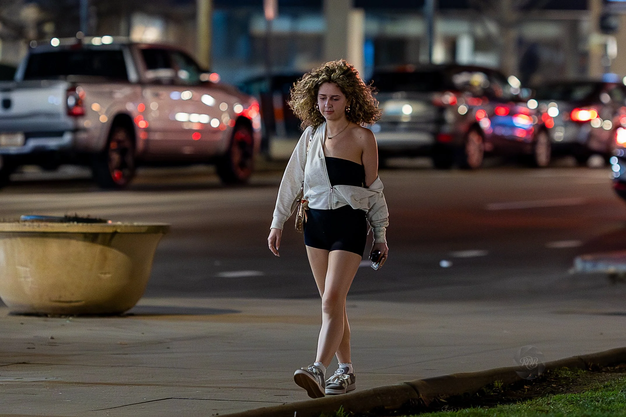 A young woman with curly hair walking on a city sidewalk at night, dressed in a black outfit with a light-colored jacket, holding a phone and a small bag, with parked cars and city lights in the background.