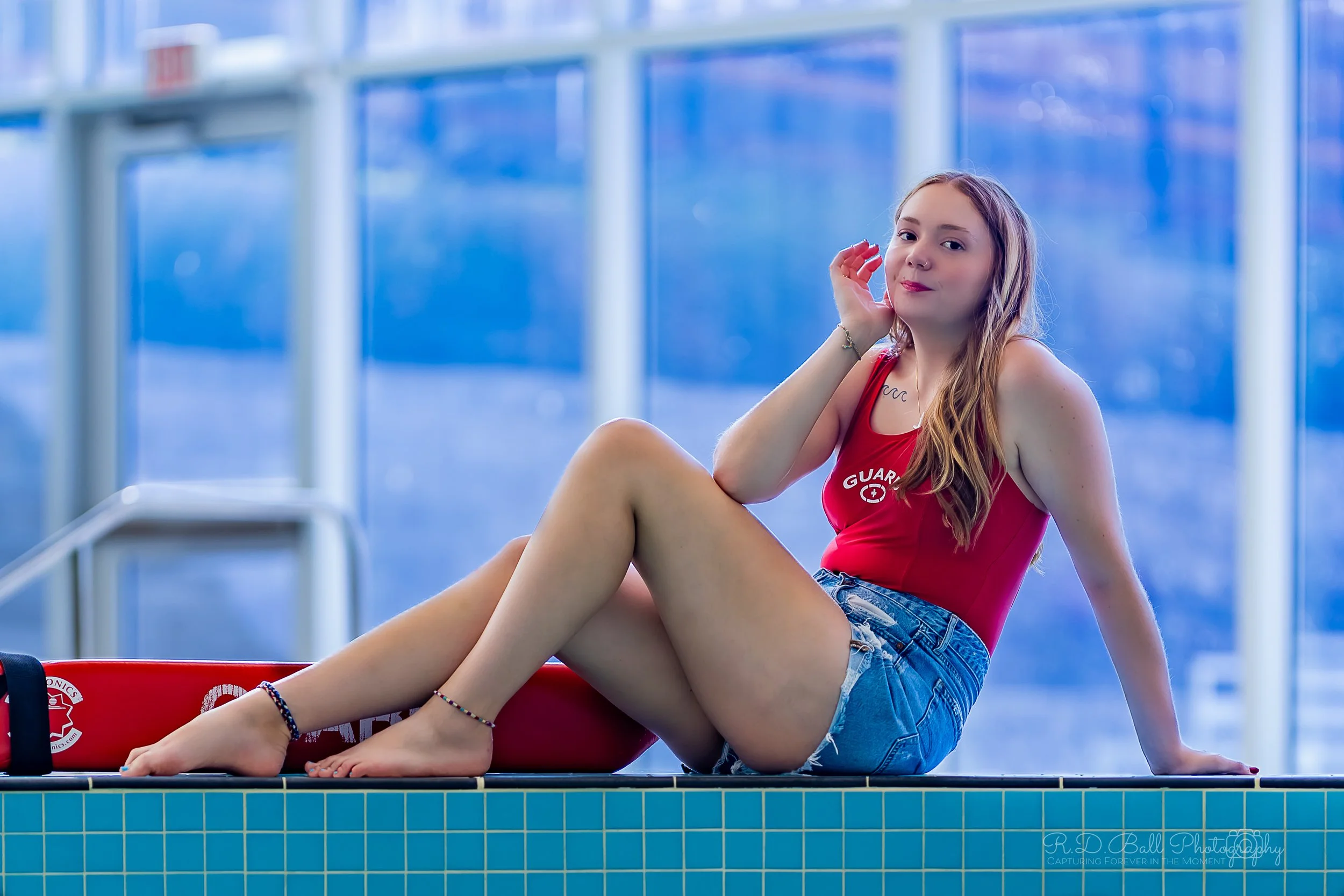 Young woman sitting on the edge of a swimming pool, wearing a red tank top and denim shorts, with a red rescue tube nearby, inside a pool area with glass windows.