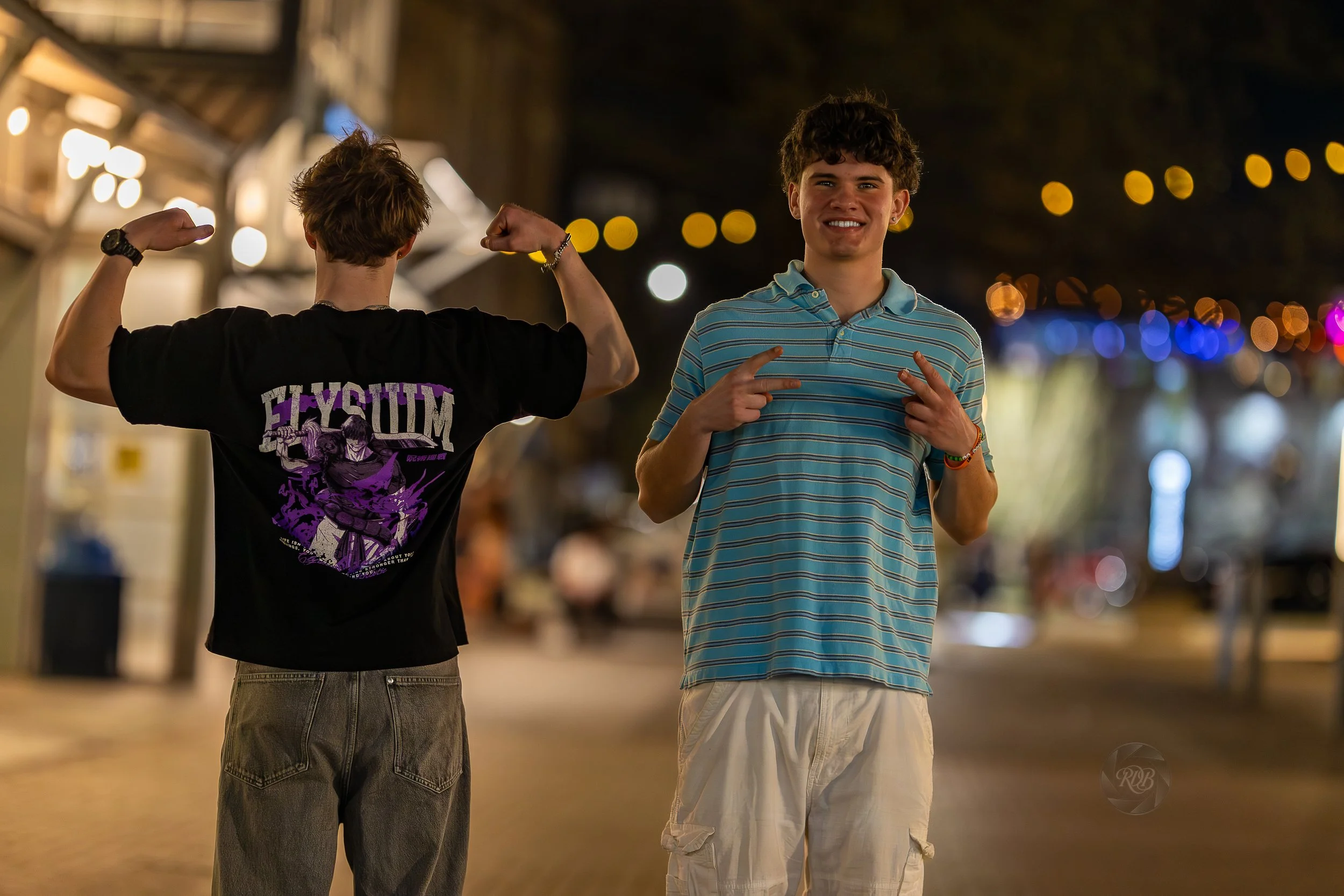 Two young men outdoors at night, one facing away with muscles flexed, wearing a black T-shirt with purple graphic, the other smiling, making finger gun gestures, wearing a blue striped polo shirt and beige shorts, illuminated by string lights in the 