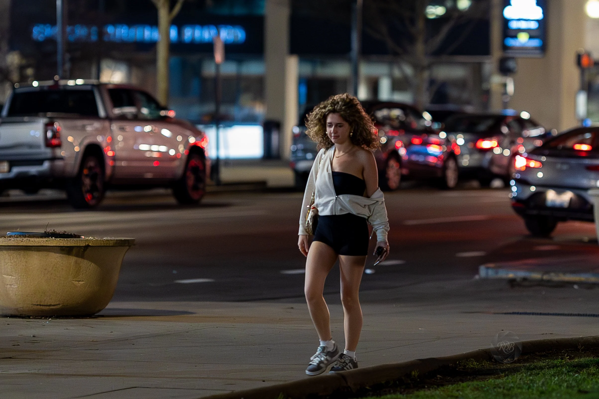A young woman walking on a city sidewalk at night, wearing a black dress, white jacket hanging off her shoulders, and sneakers, with parked cars and illuminated signs in the background.