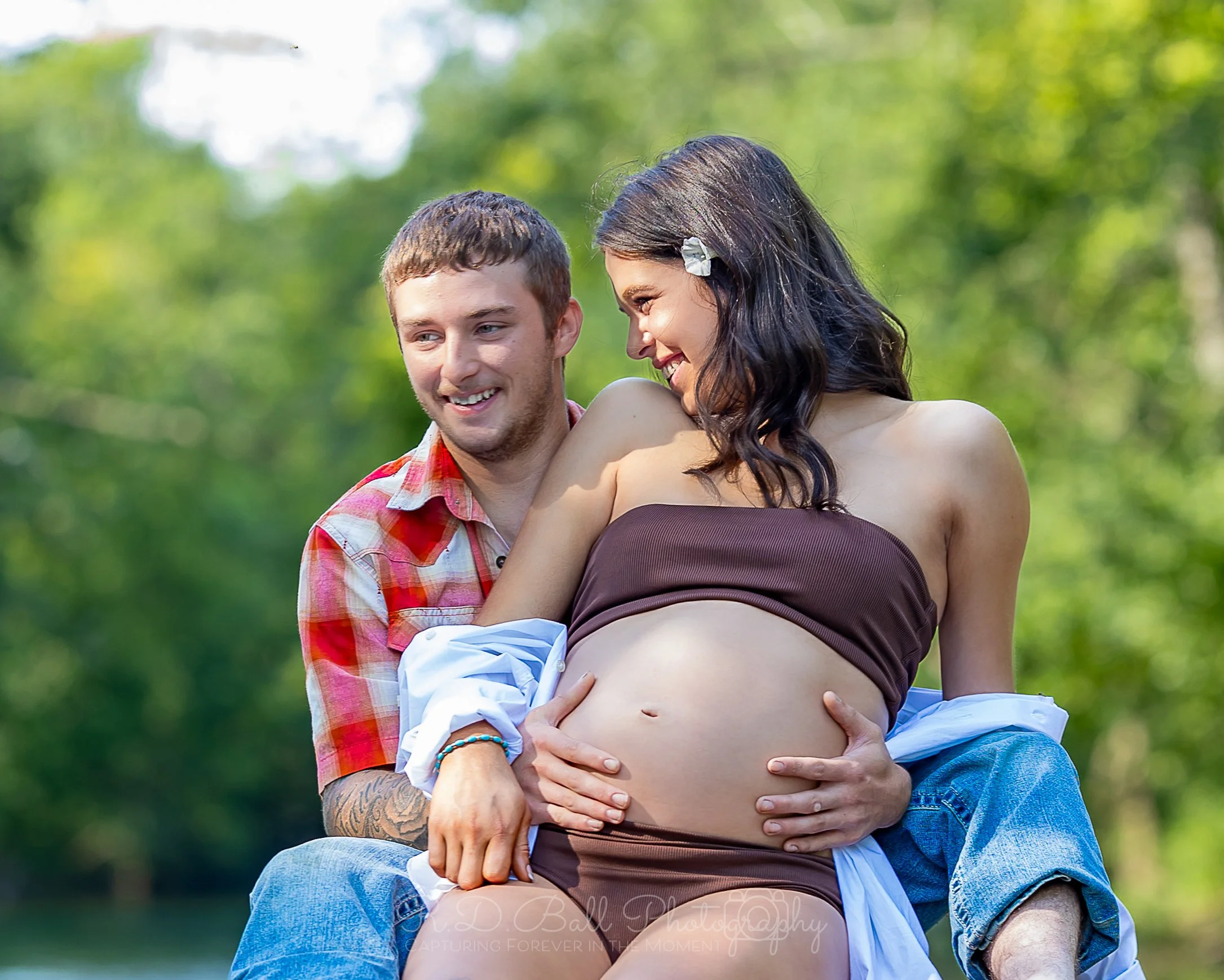A pregnant woman and a man sit outdoors, smiling and loving, with the man's hands on her pregnant belly, surrounded by greenery.