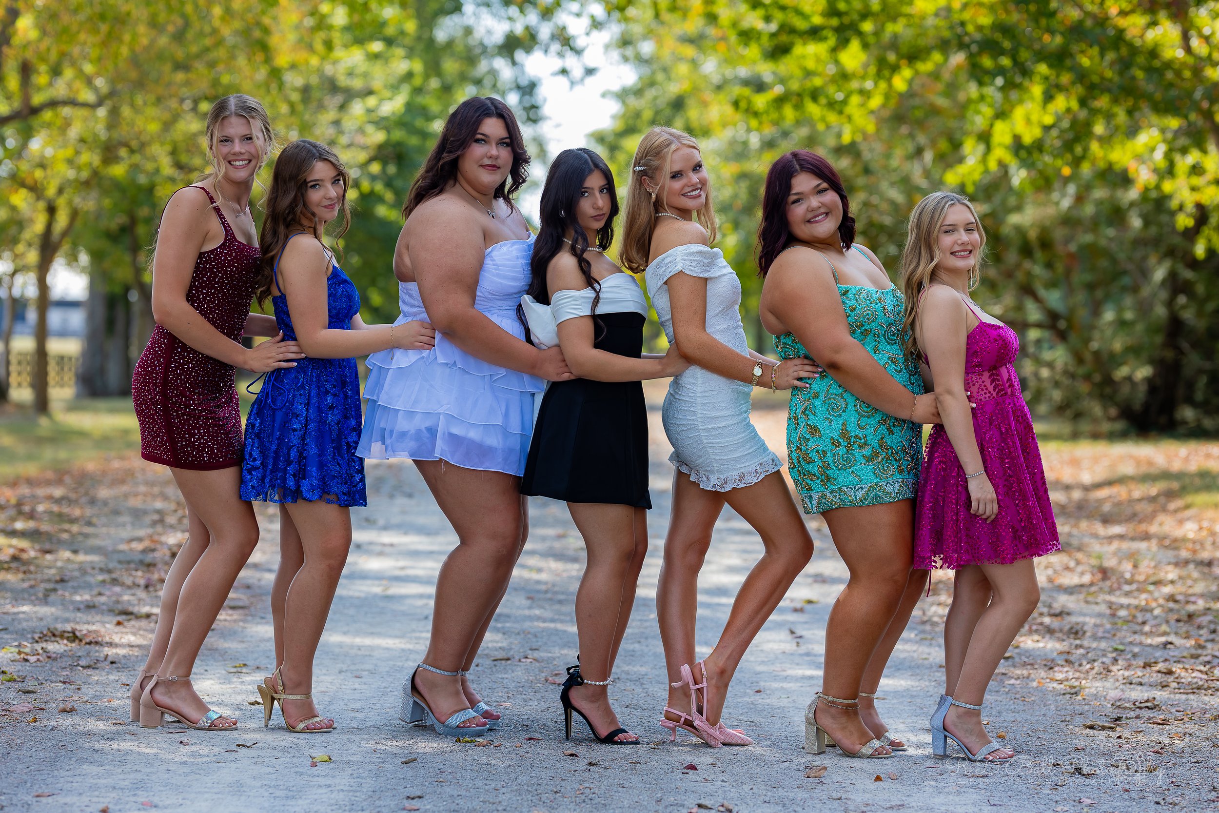 Seven women standing in a line outdoors, holding onto each other's hips, dressed in colorful dresses and high heels, with trees and a path in the background.