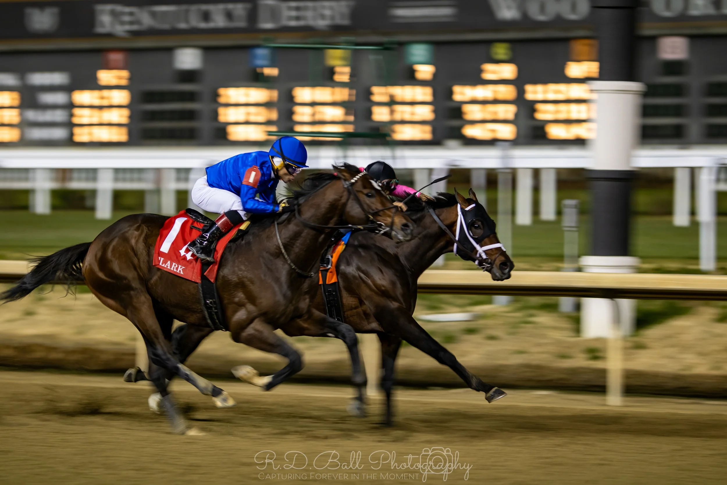 Horse race with two jockeys racing on a dirt track at night, scoreboard in the background.
