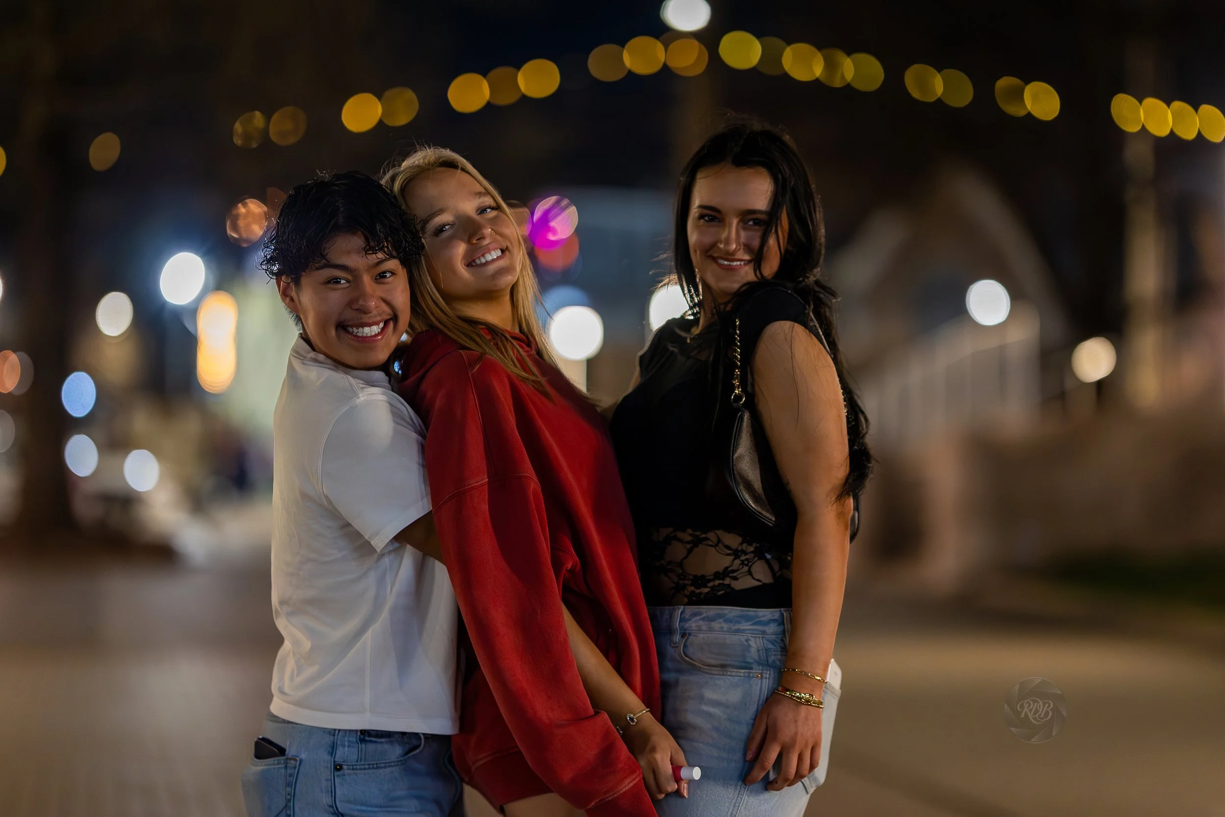 Three young women smiling together at night with blurred city lights in the background.
