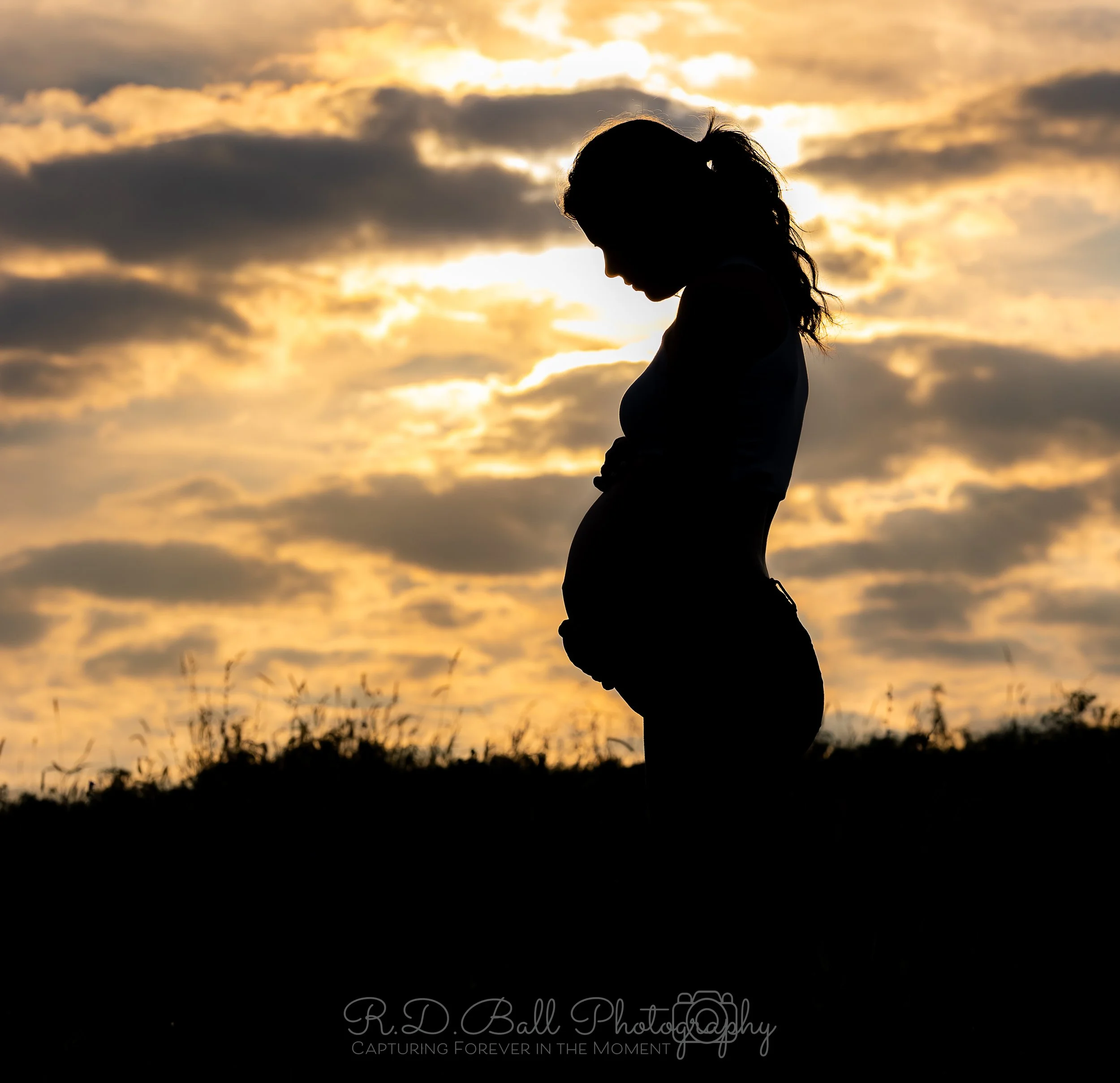 Silhouette of a pregnant woman standing outdoors during sunset with clouds in the sky.
