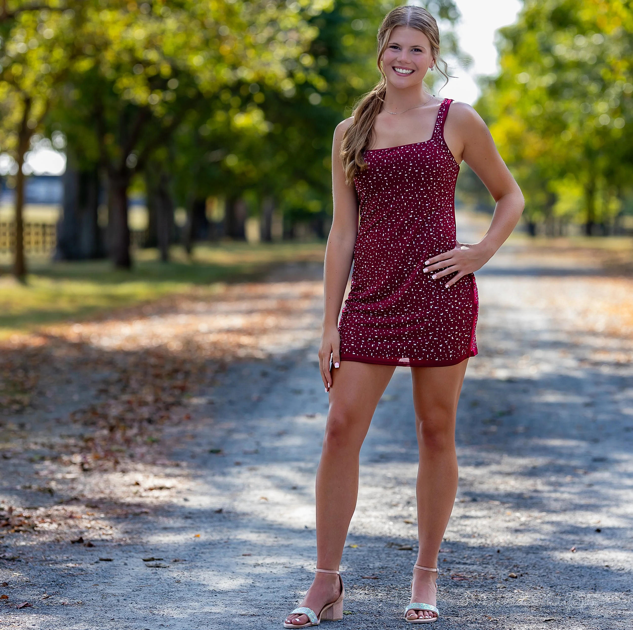 A young woman in a red polka-dot dress standing on a dirt path in a park with trees and sunlight in the background.