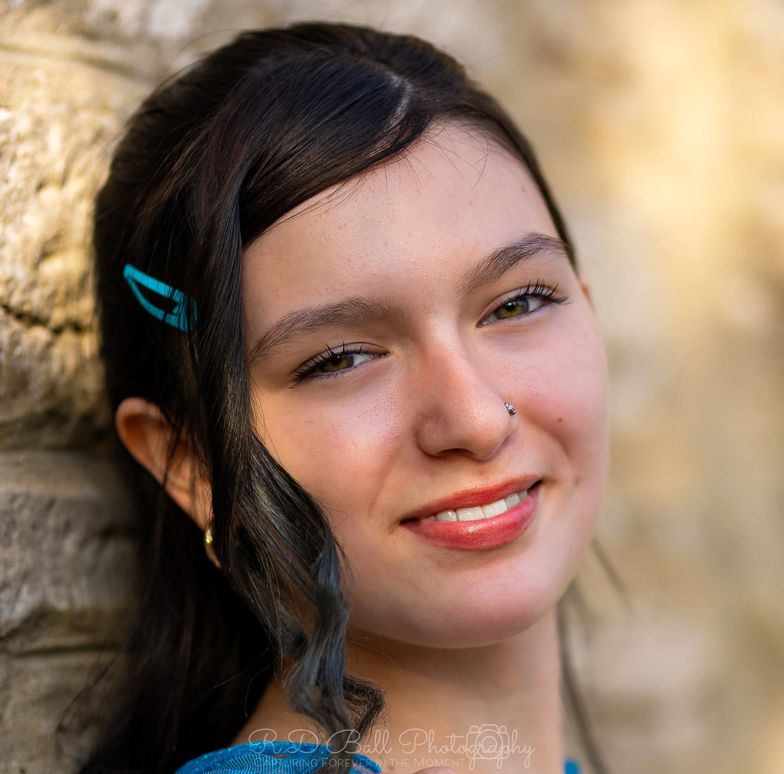 Close-up of a young woman with dark hair, a blue hair clip, and a nose piercing, smiling while leaning against a stone wall.