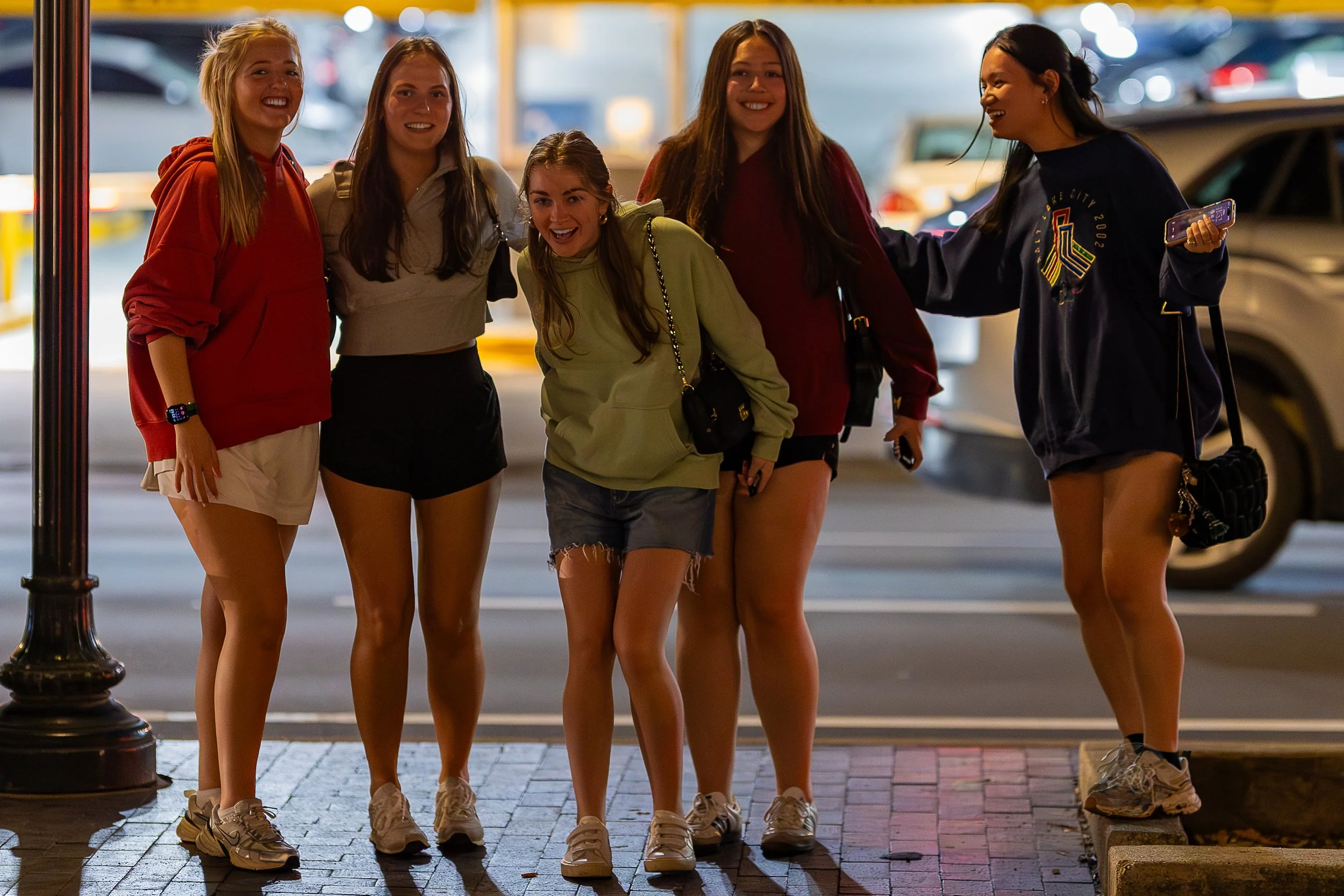 Five young women standing on a city street at night, smiling and posing for a photo together, with cars in the background.
