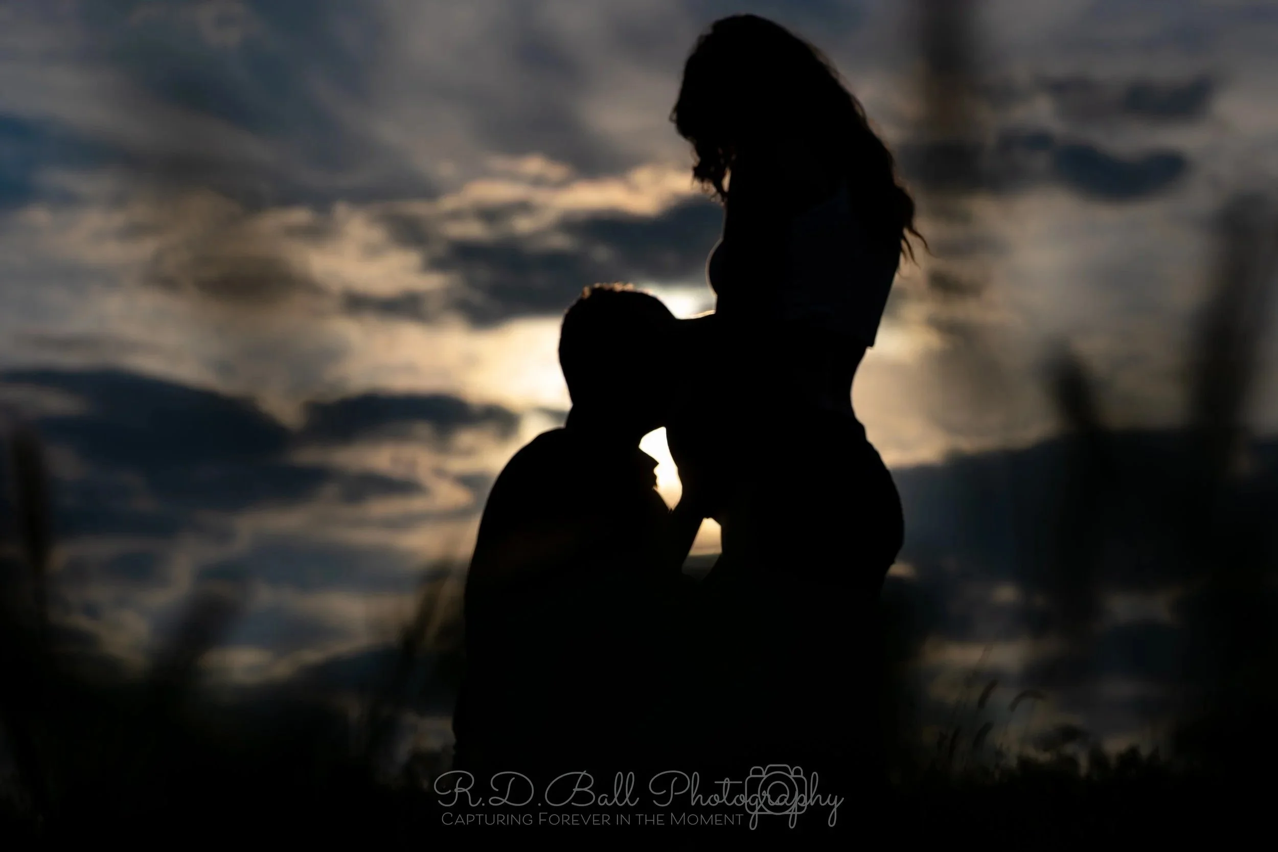 Silhouetted couple, with the man kneeling and the woman standing, touching foreheads against a sunset sky with clouds.