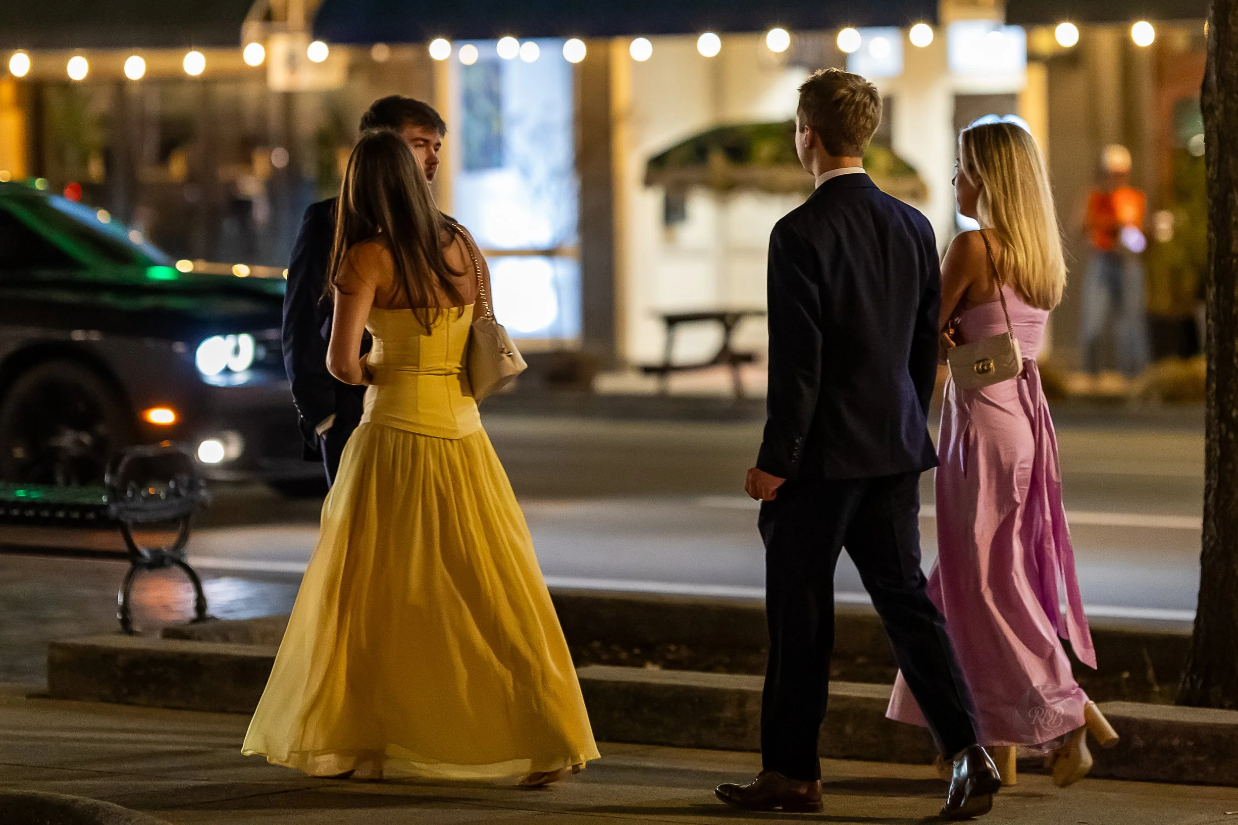 Four people dressed in formal evening attire conversing on a city sidewalk at night, illuminated by string lights.