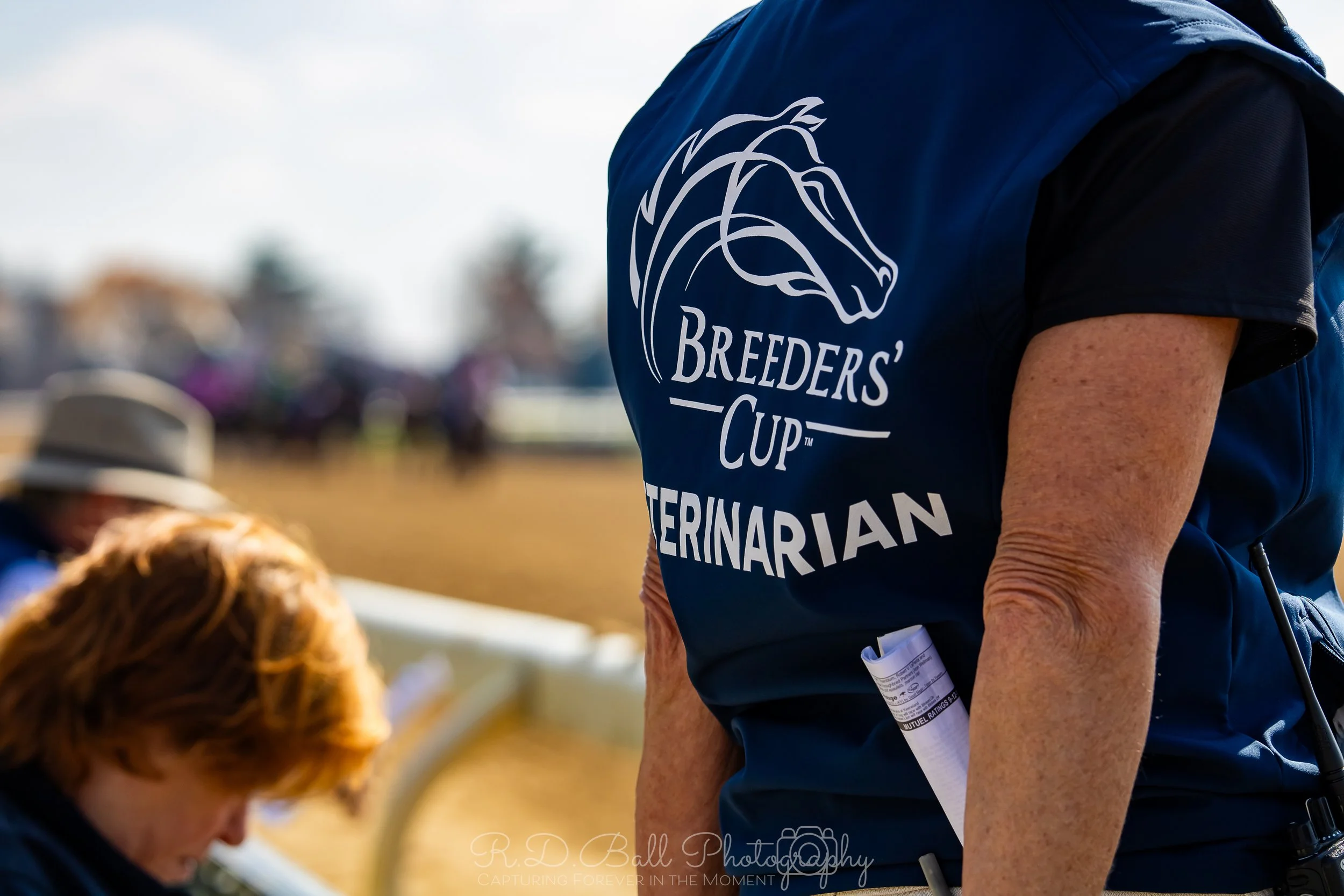 Close-up of a person wearing a navy blue vest with a logo for 'Breeders' Cup Veterinarian' on the back, holding a rolled-up paper, during an outdoor event.