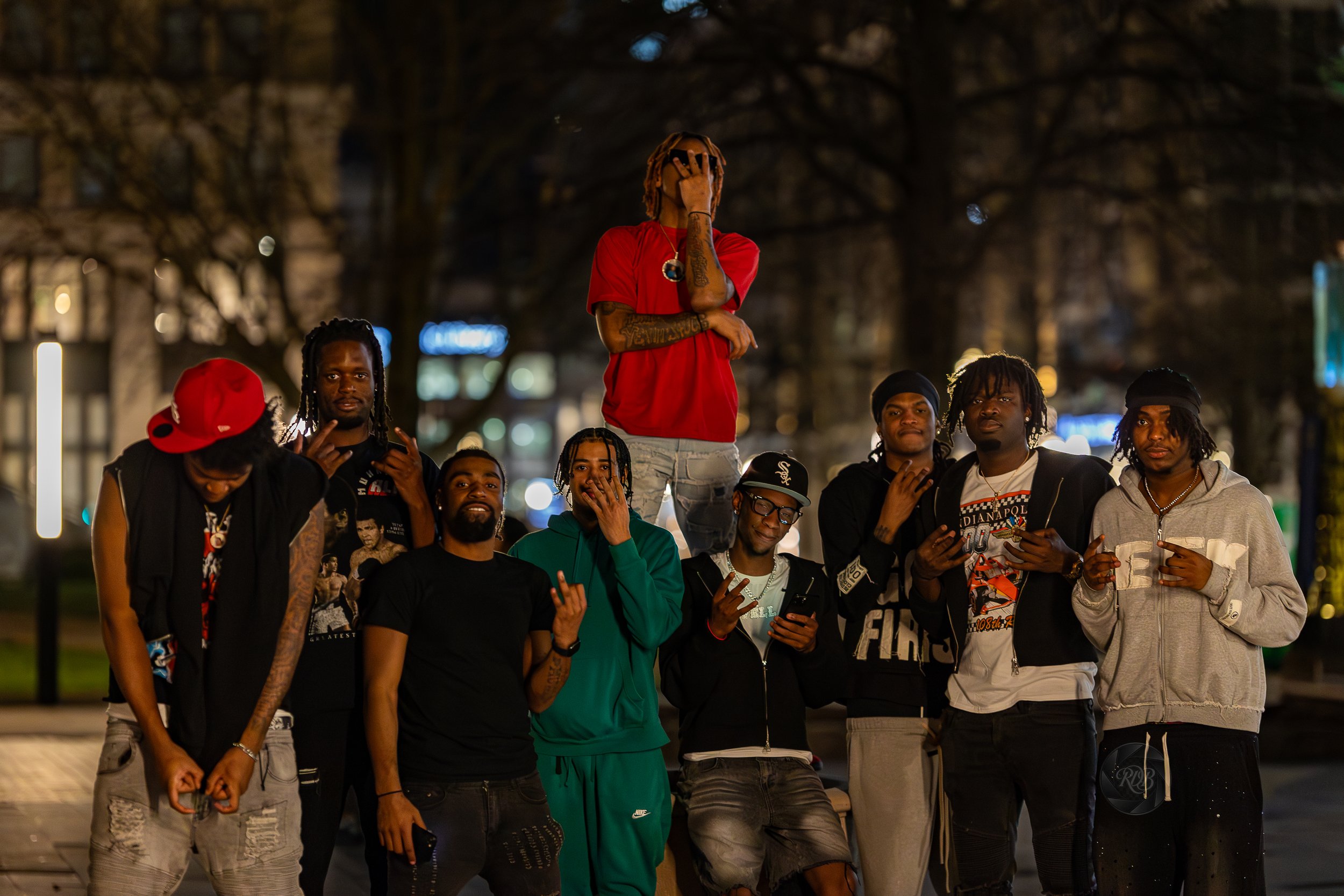 A group of ten young men gathered outdoors at night, with one man standing on a ledge or bench in the center. The group is casually dressed, some with tattoos, wearing hoodies, t-shirts, and caps. The background shows blurred city lights and trees.