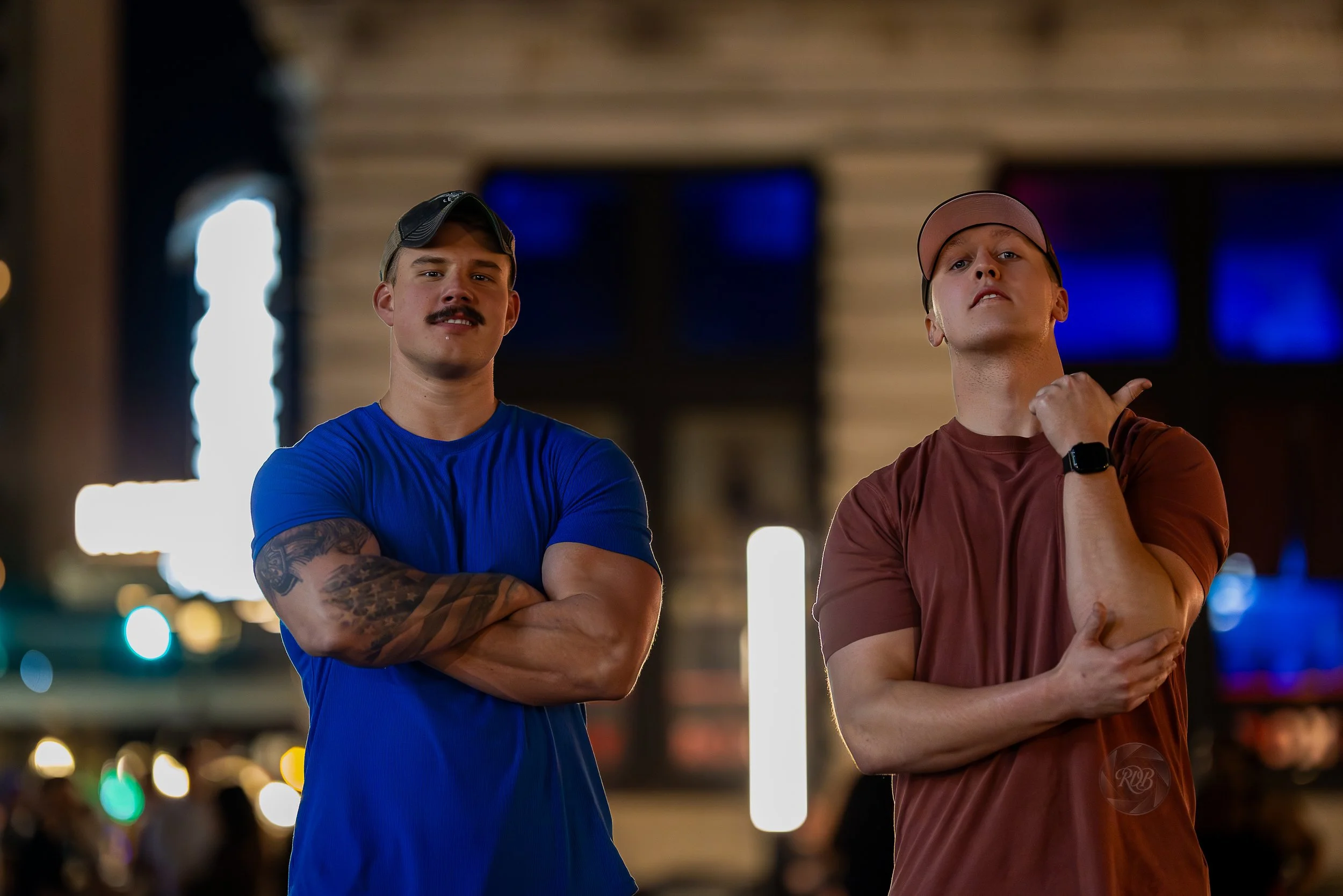 Two young men standing outdoors at night, one in a blue shirt with crossed arms and tattoos, and the other in a red shirt with a baseball cap, touching his neck, with blurred city lights in background.