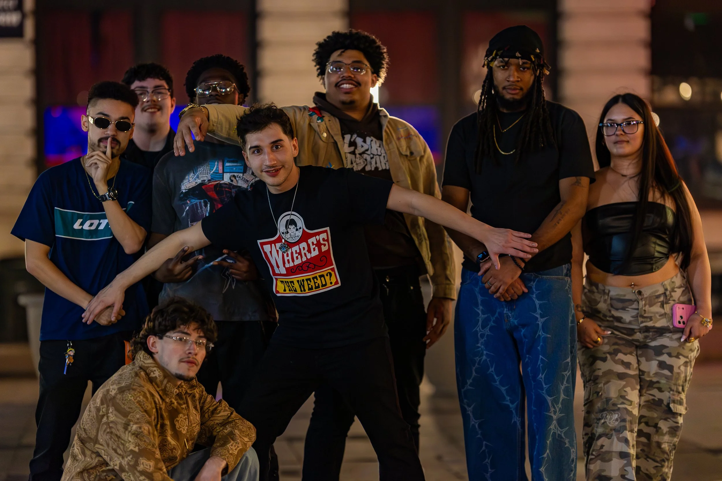 A diverse group of nine young people posing together outdoors at night, some smiling and making playful gestures, with a blurred background of warm lighting and wooden structures.