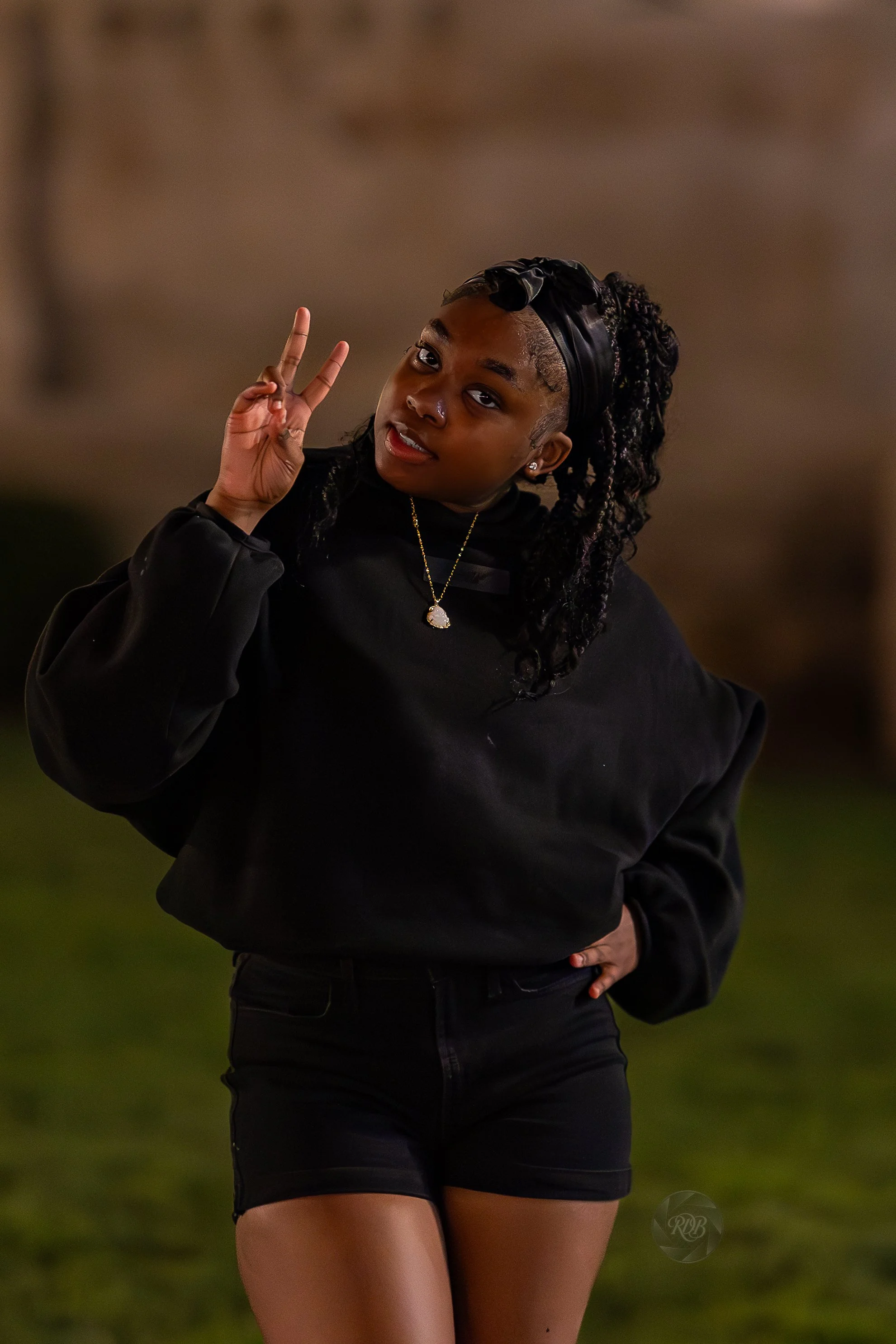 A young woman with curly hair, dressed in a black sweatshirt and shorts, making a peace sign and smiling at the camera during evening outdoors.