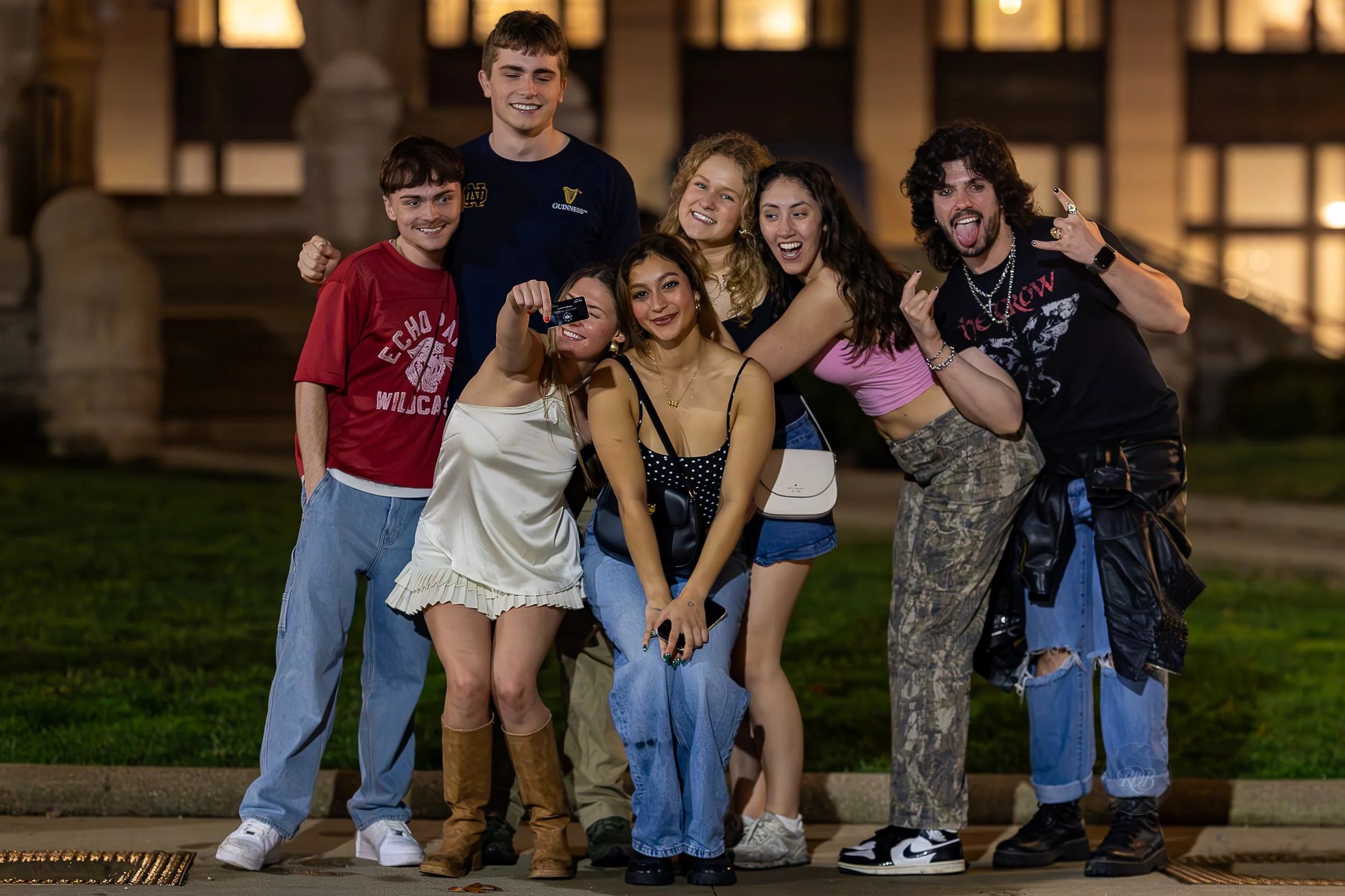 Group of seven young people posing together outside at night, smiling and making playful gestures.