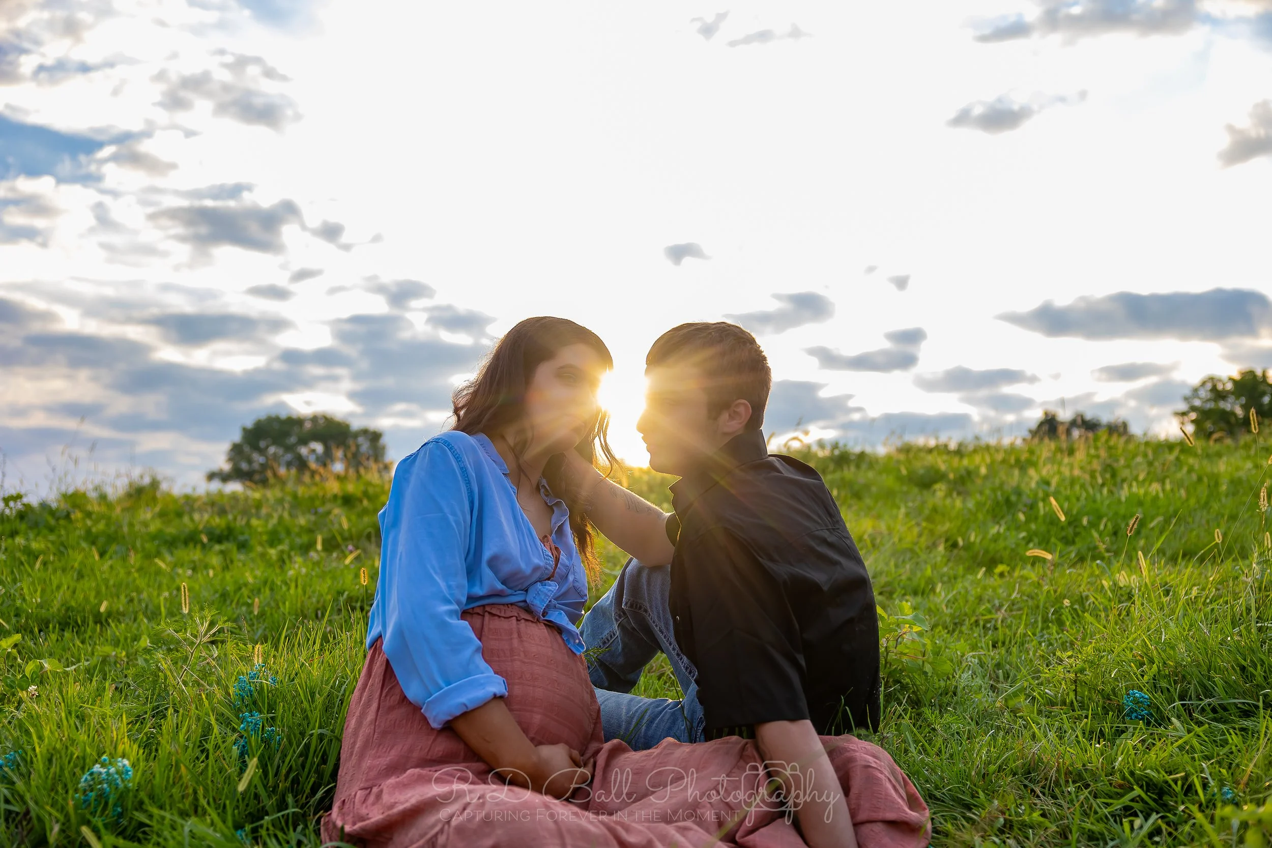 A couple sitting on a grassy field during sunset, facing each other with sunlight shining between them, blue sky with clouds behind them.