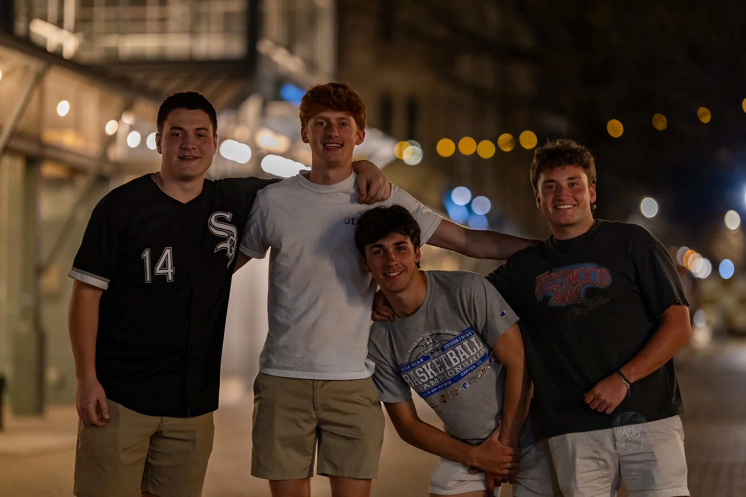 Four young men smiling and posing with their arms around each other at night, with blurred lights in the background.