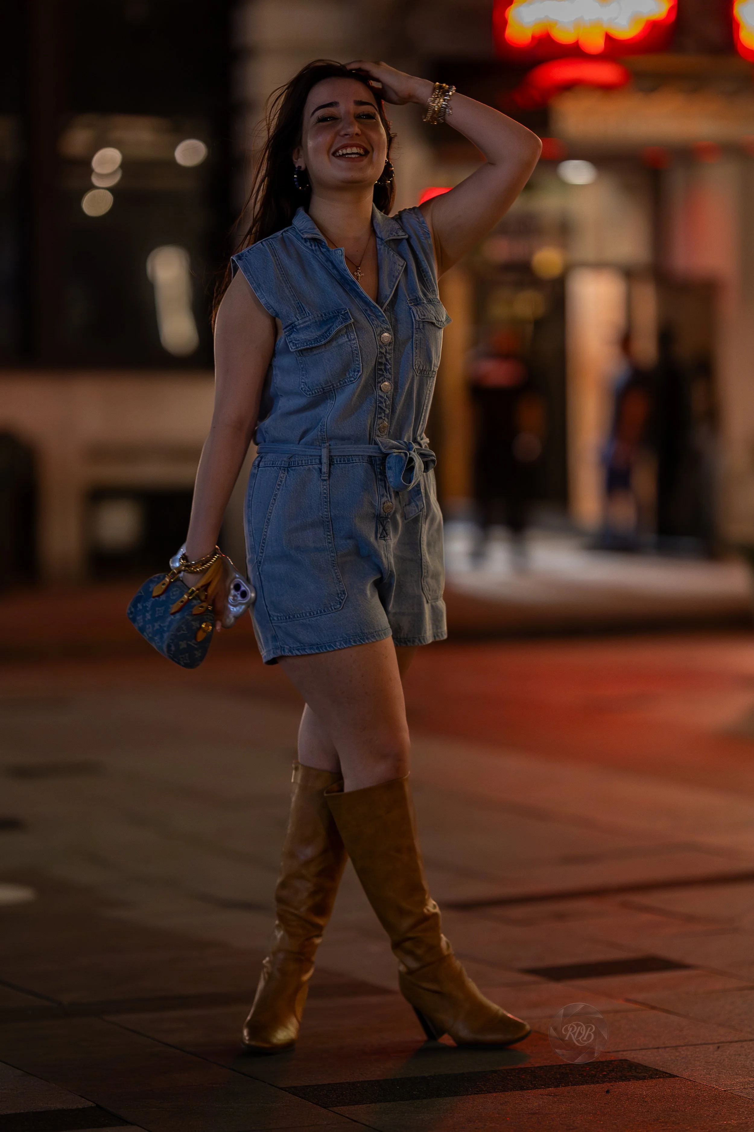 A woman with long dark hair in a denim romper with rolled sleeves and matching shorts, smiling and posing on a city street at night. She is wearing tall tan leather boots, gold jewelry, and holding a Louis Vuitton handbag.