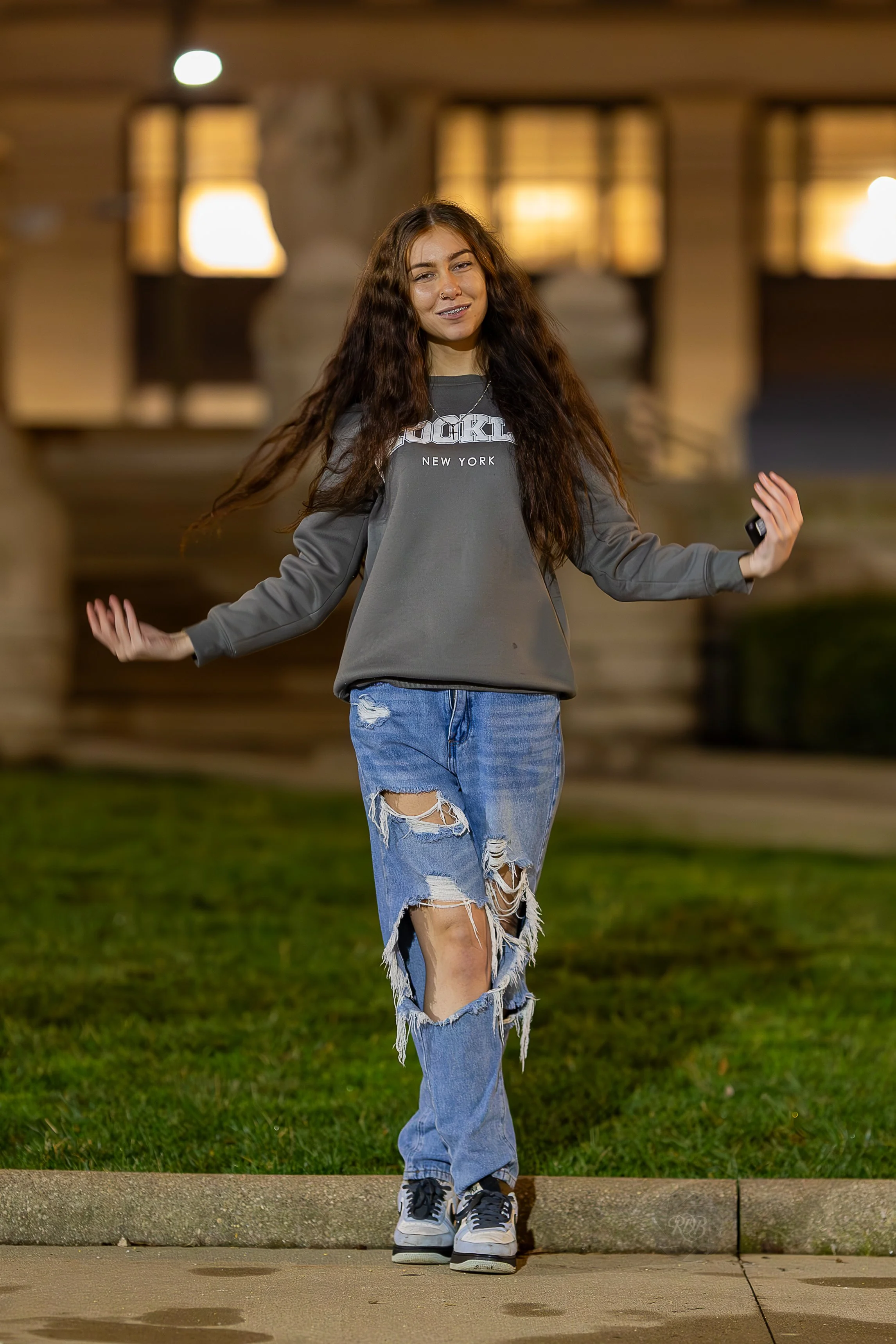 A young woman with long, curly hair standing outdoors at night on a sidewalk in front of a well-lit building, wearing a gray sweatshirt with 'Brooklyn New York' printed on it, distressed ripped jeans, and sneakers.