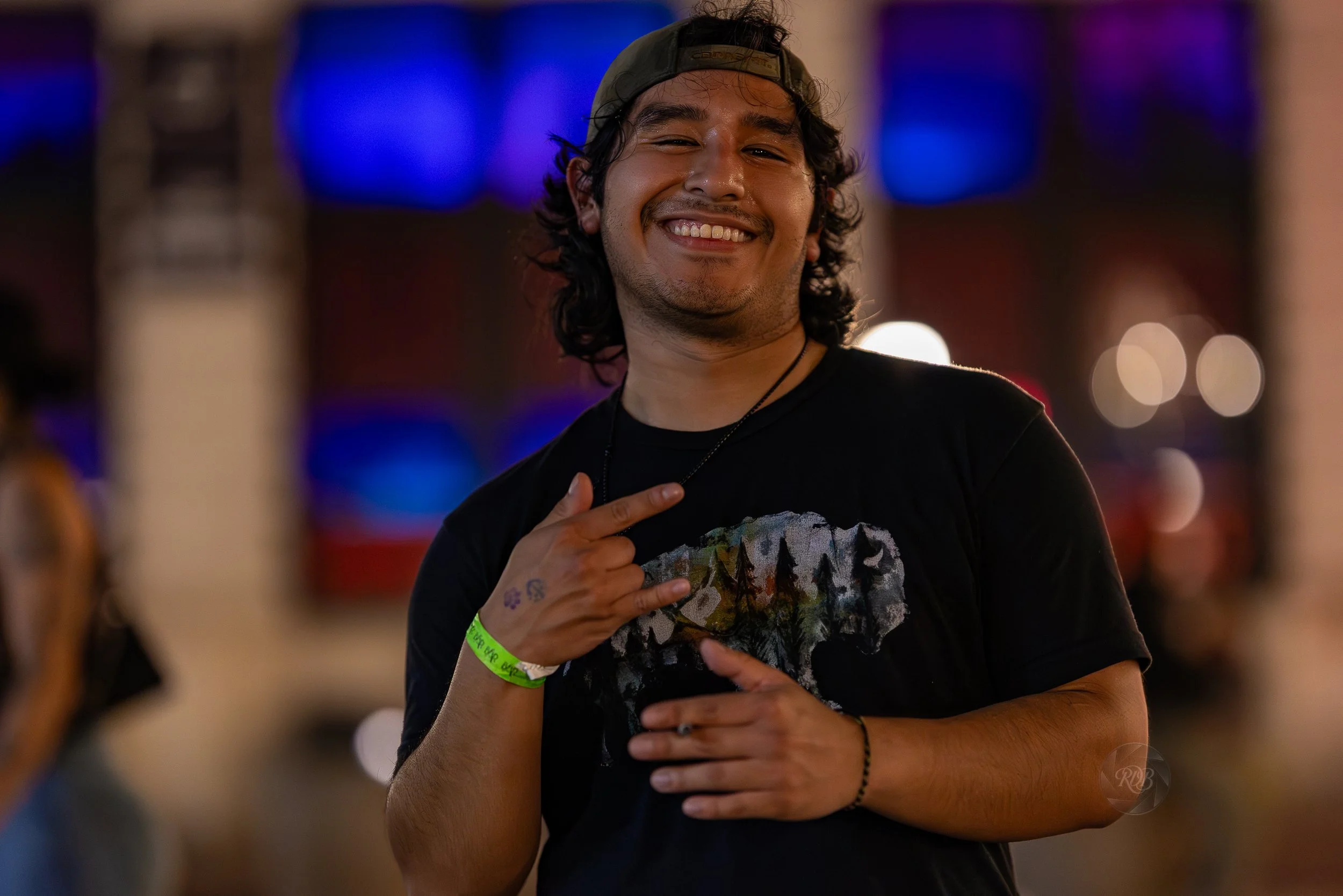 A young man smiling and making a rock and roll hand gesture at a lively event or party, with colorful lights and blurred background.