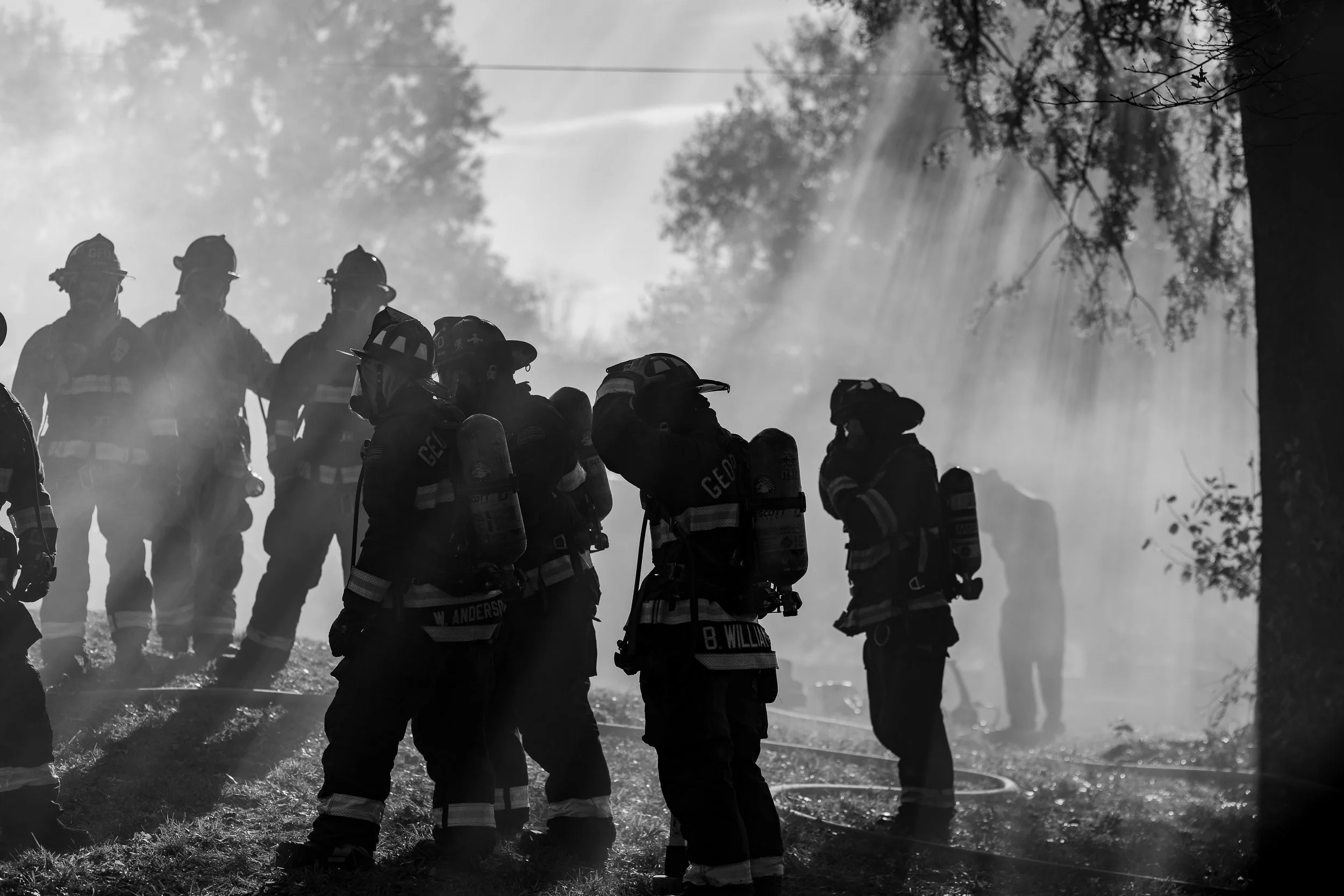 Silhouettes of firefighters in firefighting gear standing amidst smoke and rays of light.