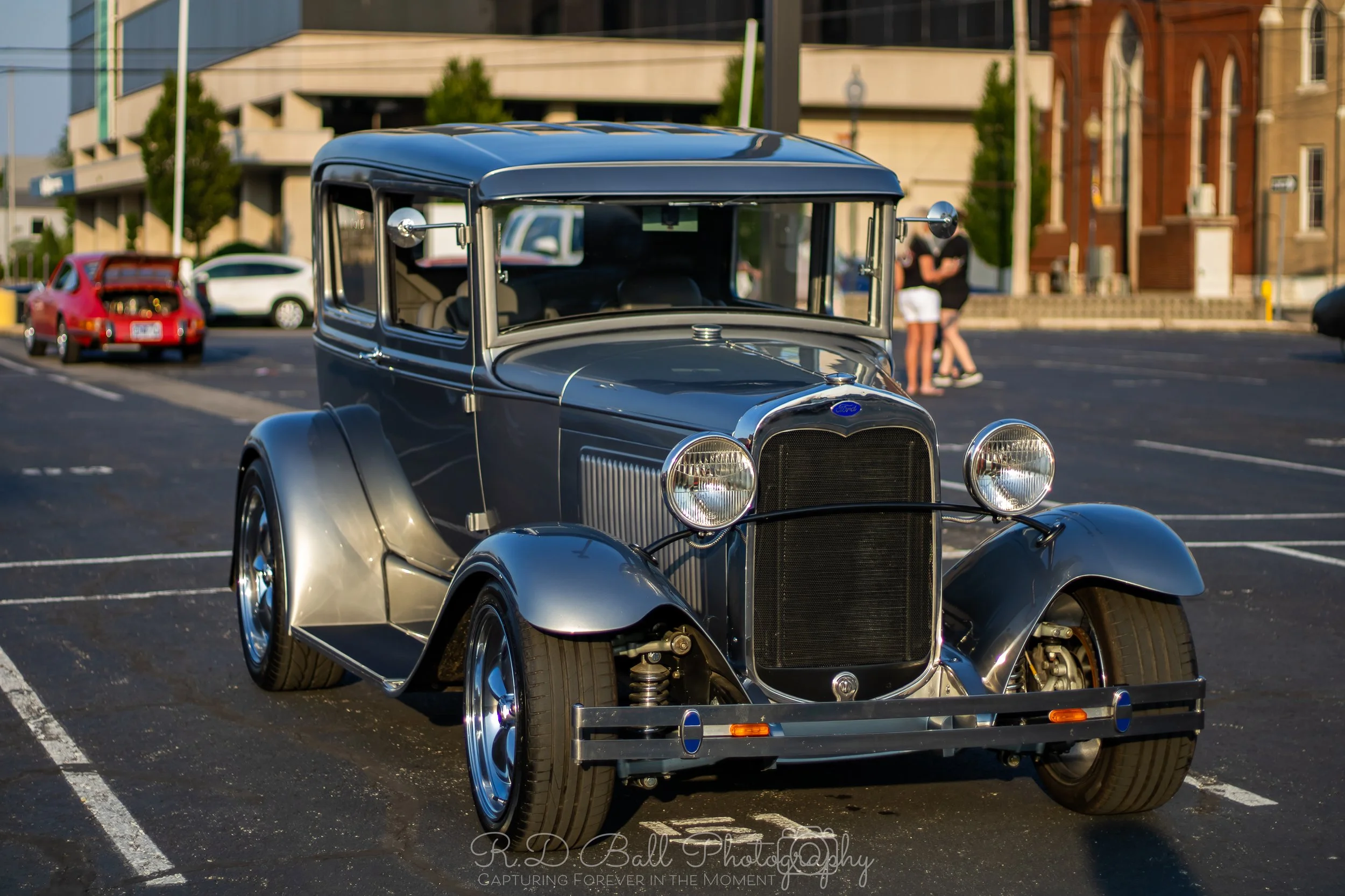 Vintage black car parked in a parking lot with a yellow building and modern structures in the background.