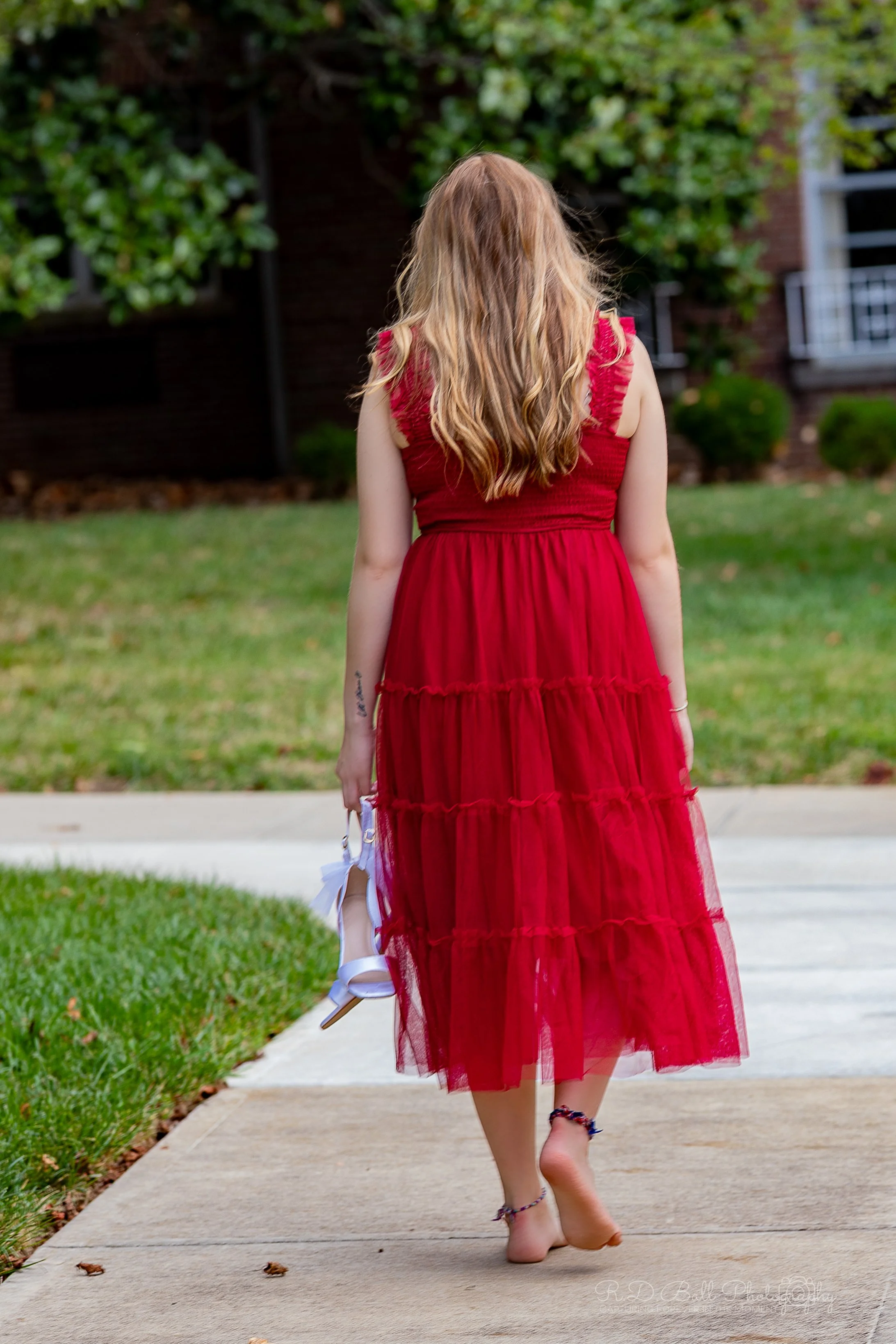 A woman with long, wavy blonde hair walking barefoot on a sidewalk, wearing a red dress and holding white high-heeled shoes in her right hand.