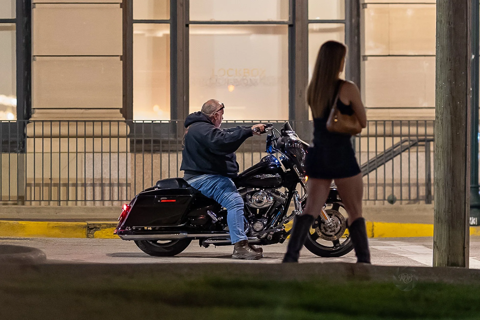 A man in a black hoodie and jeans sitting on a black motorcycle on the street at night, with a young woman in a black dress and boots walking past him, holding a brown bag, in front of a building with large windows.